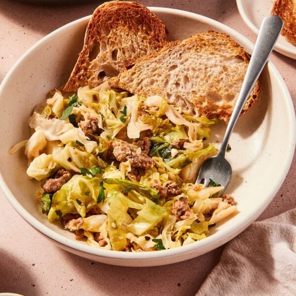 Large white bowl filled with serving of skillet sausage and cabbage with slices of toasted bread also in bowl with fork picking up a bite of cabbage.
