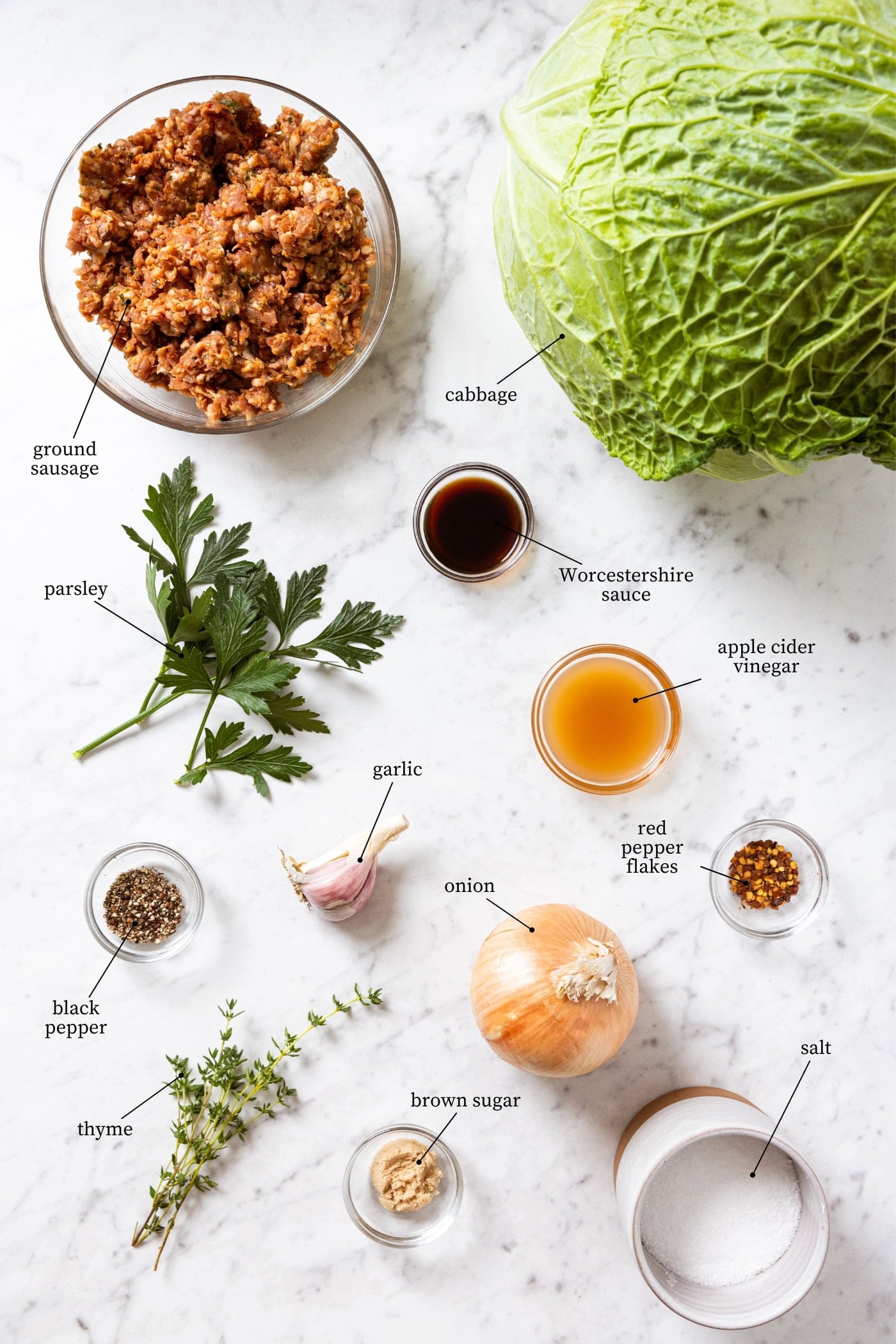 White marble surface filled with all ingredients needed to make skillet sausage and cabbage recipe including a whole cabbage, ground sausage, herbs, garlic, onion, spices, and more.