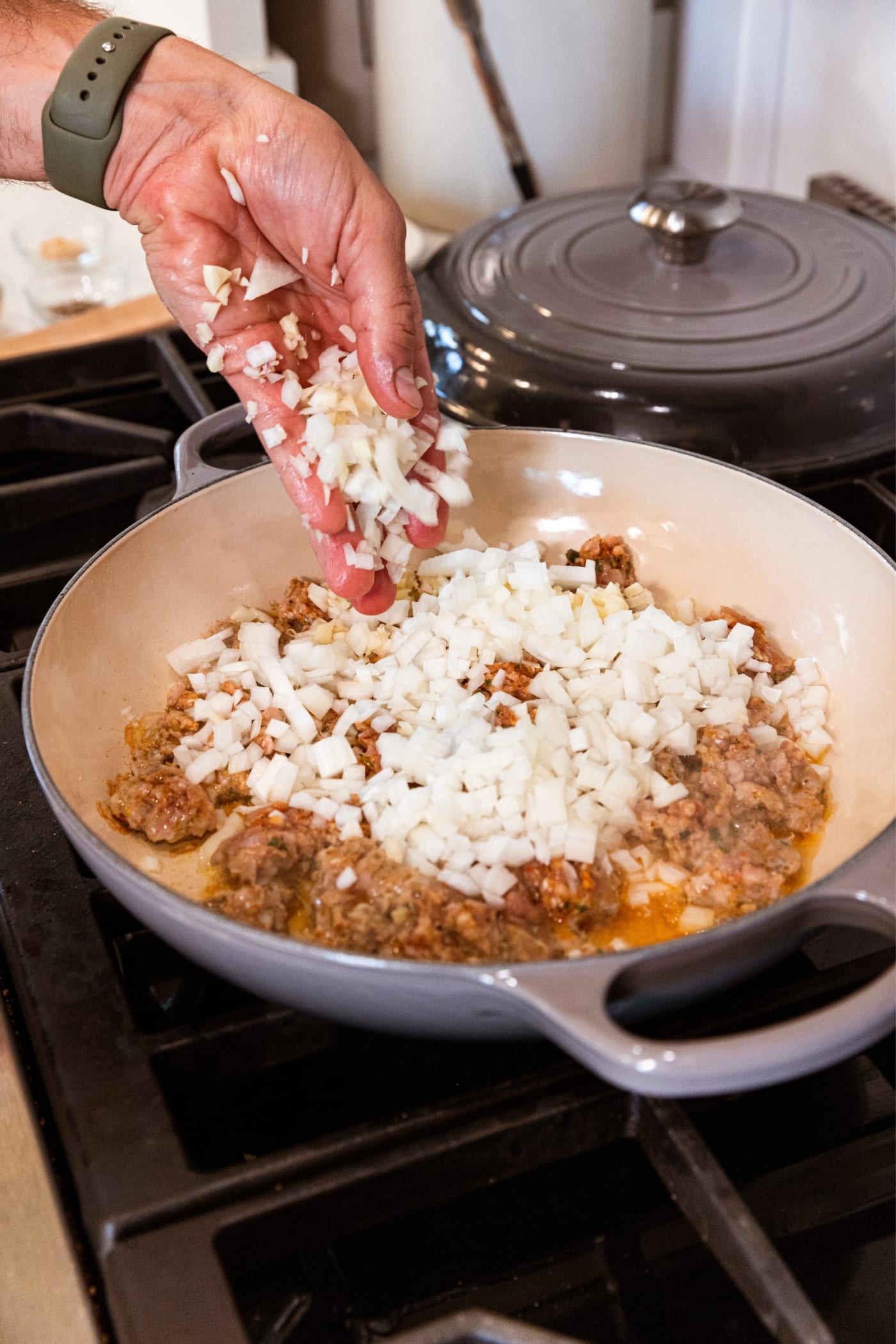 Hand pouring diced onion into large gray skillet on stovetop with cooking ground sausage underneath.