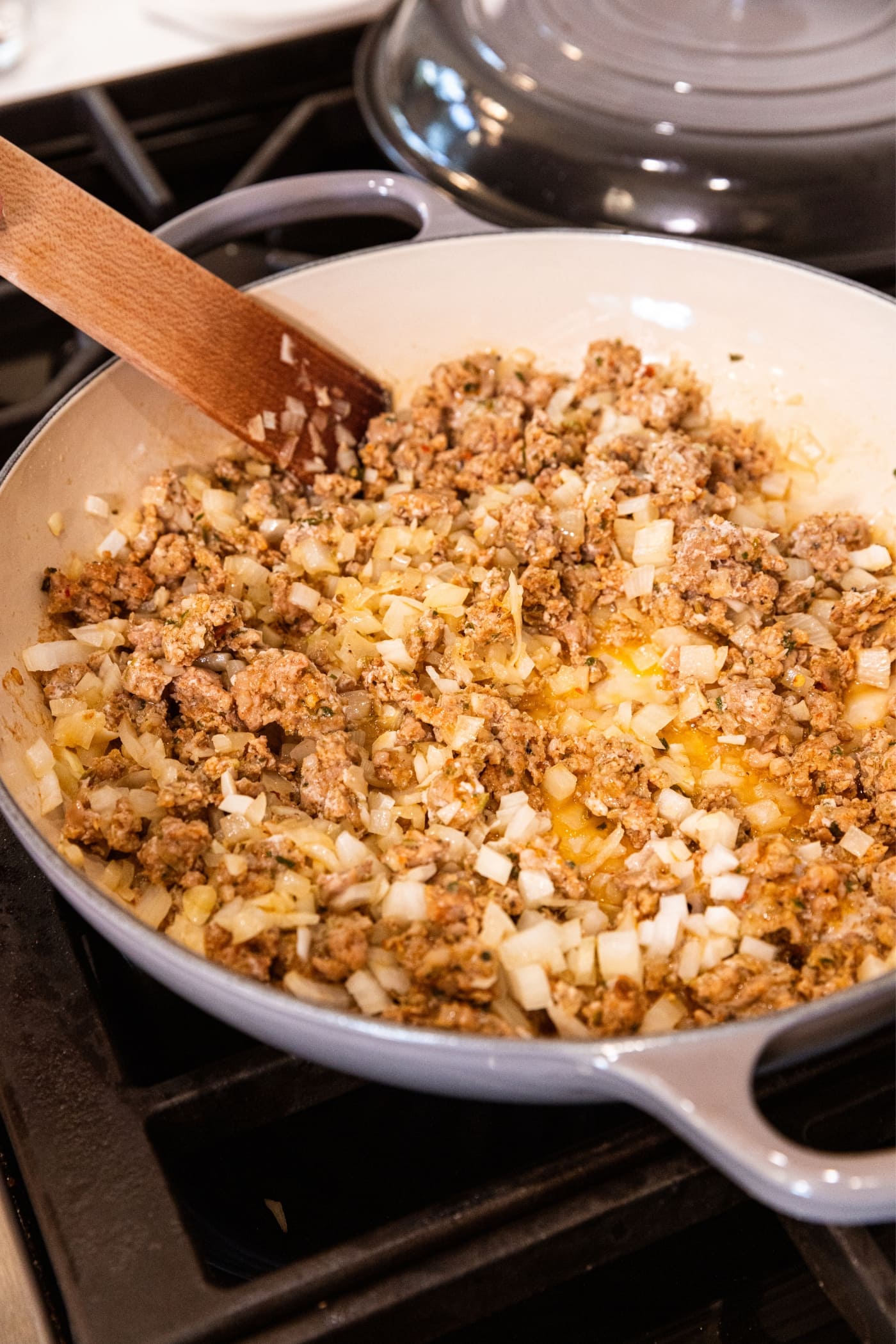 Wooden spoon mixing together ground sausage, onion, and minced garlic mixture in large gray skillet on the stovetop.