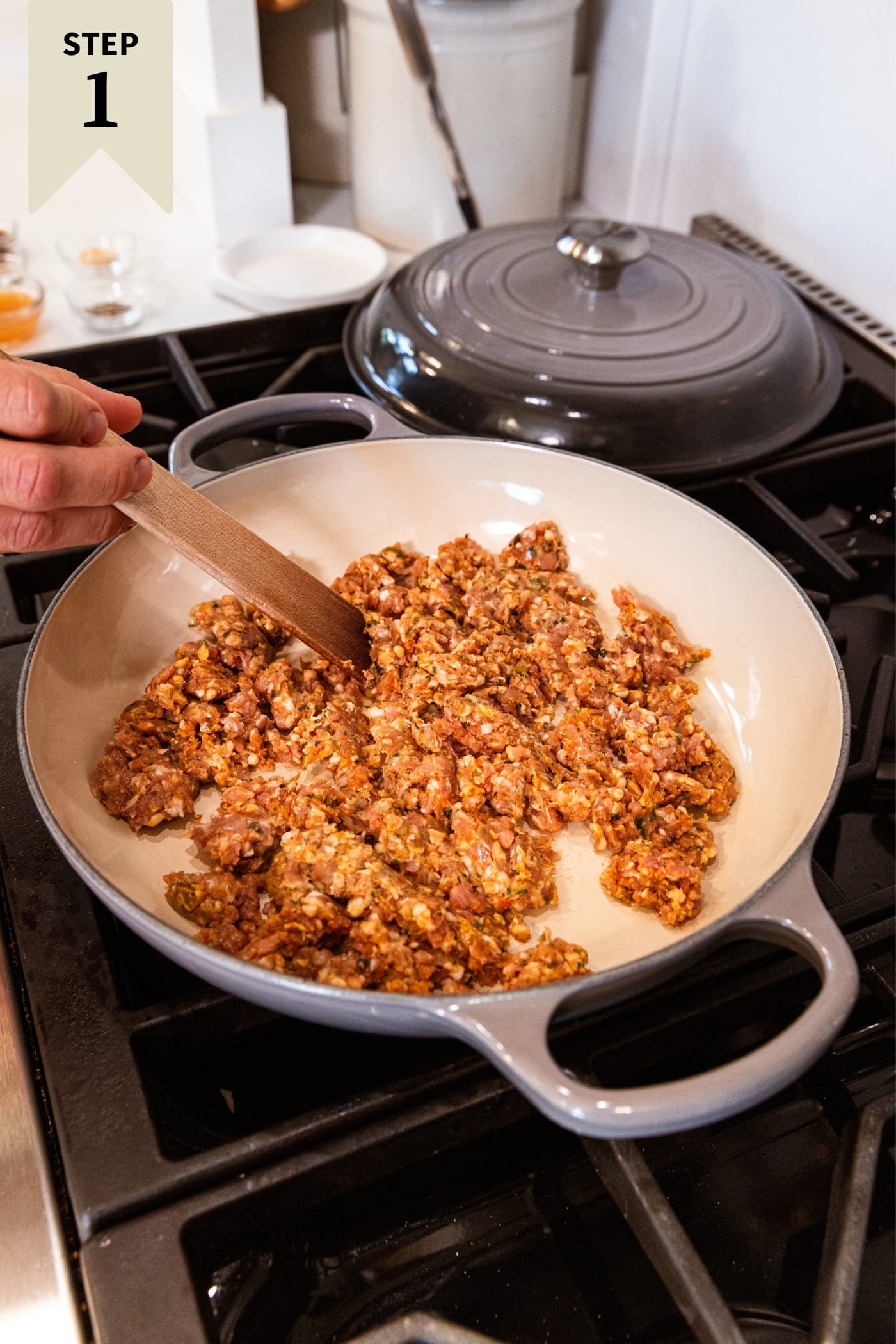 Hand holding wooden spoon breaking up ground sausage in large skillet on stovetop with slight browning.