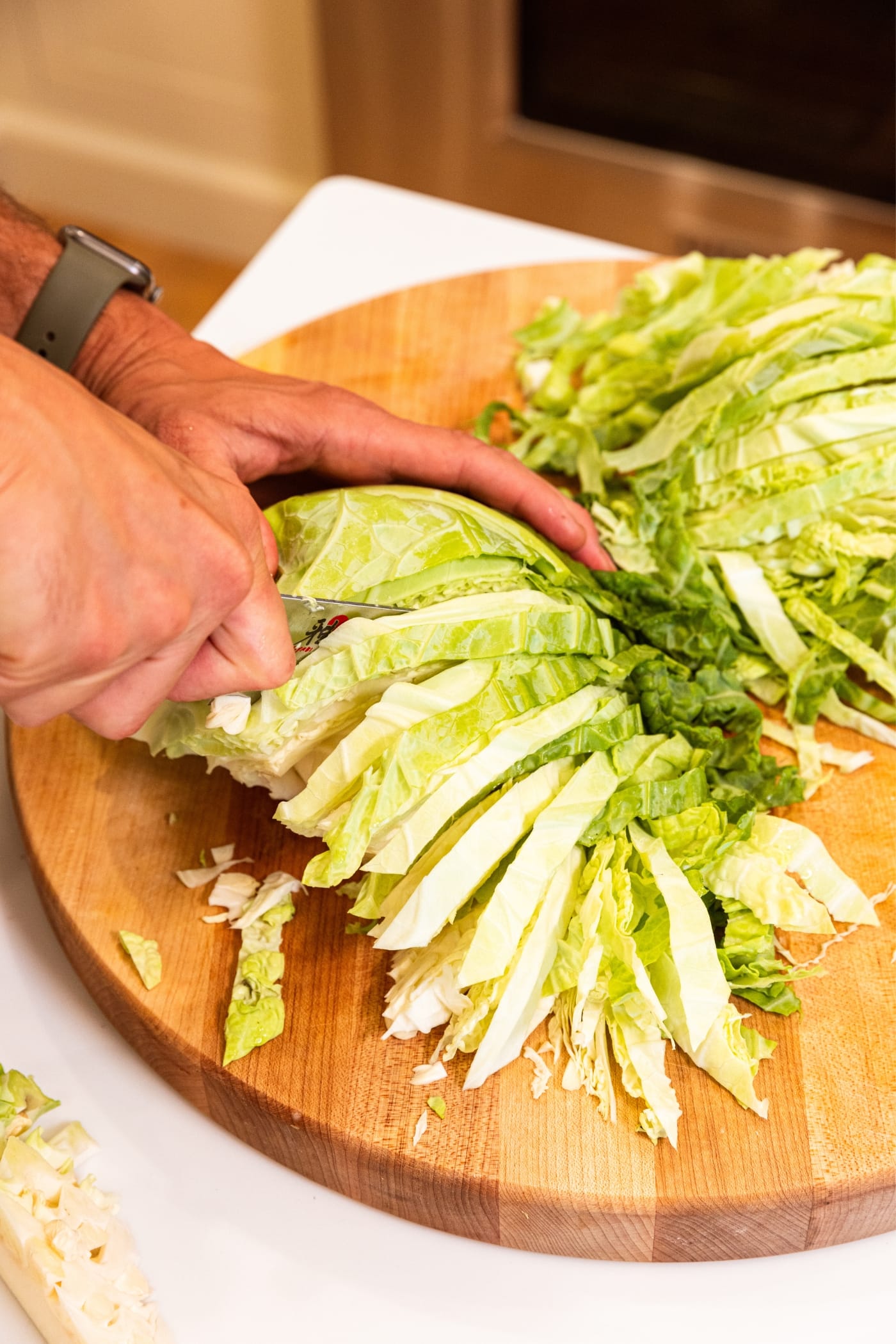 Hand using large knife to slice light green colored cabbage into small slices on wooden cutting board.