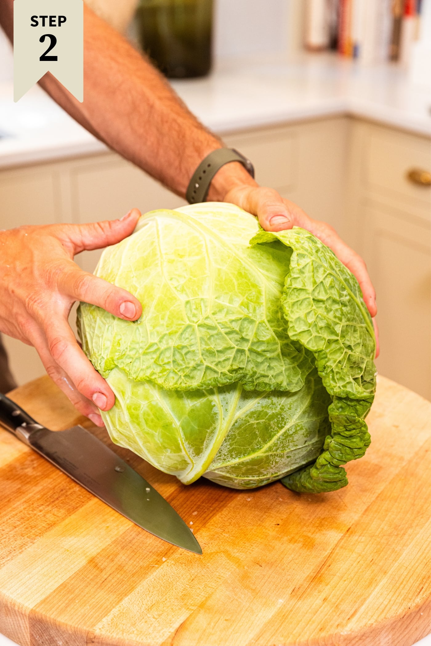 Hand holding large whole light green colored cabbage on wooden cutting board before slicing with knife.