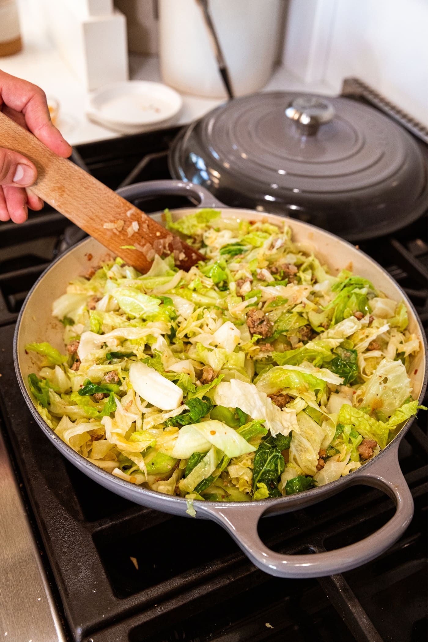 Hand holding wooden spoon stirring together cooked ground sausage with sliced cabbage on top.
