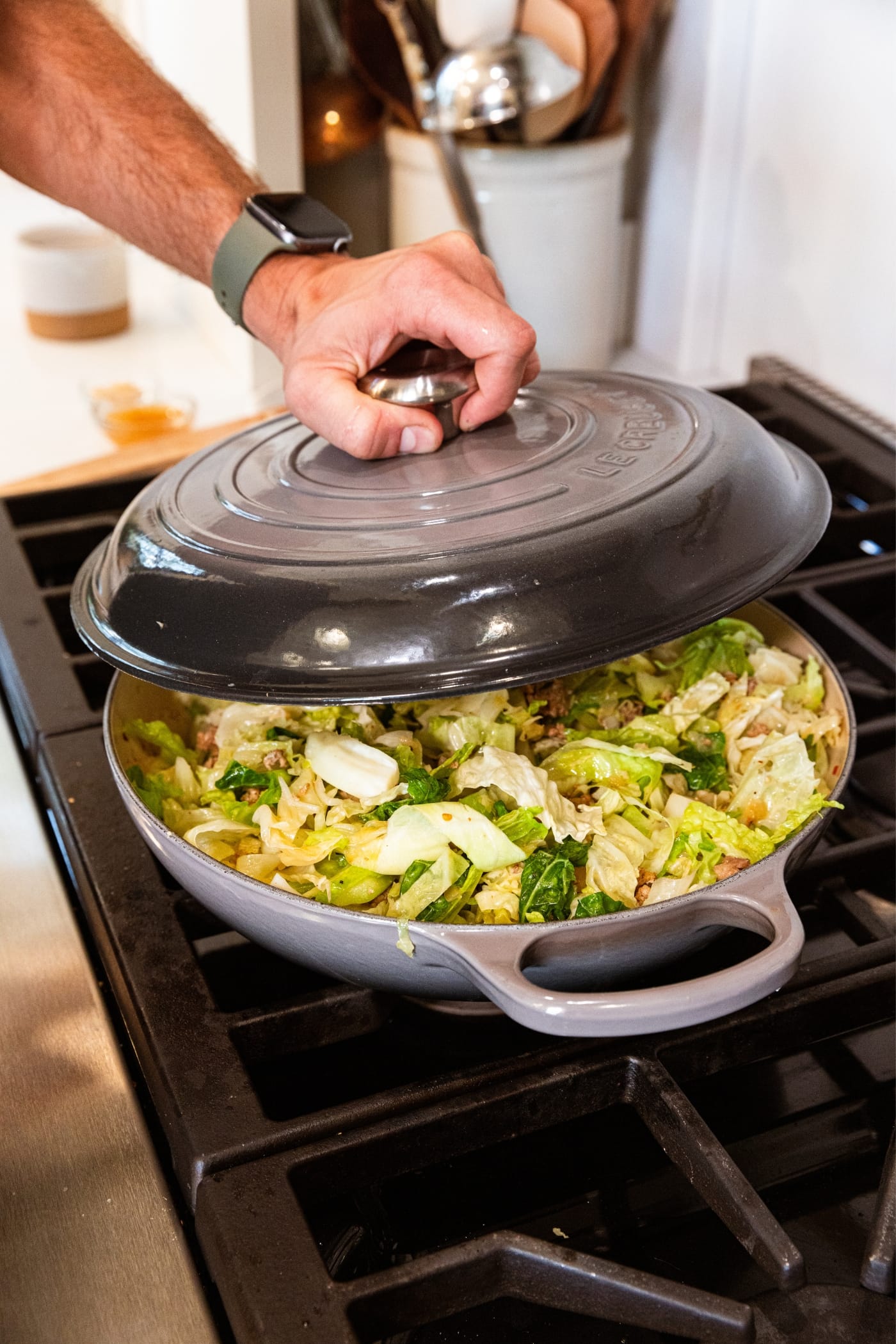 Hand holding lid of gray skillet about to place it on top of skillet filled with cooking sliced cabbage.