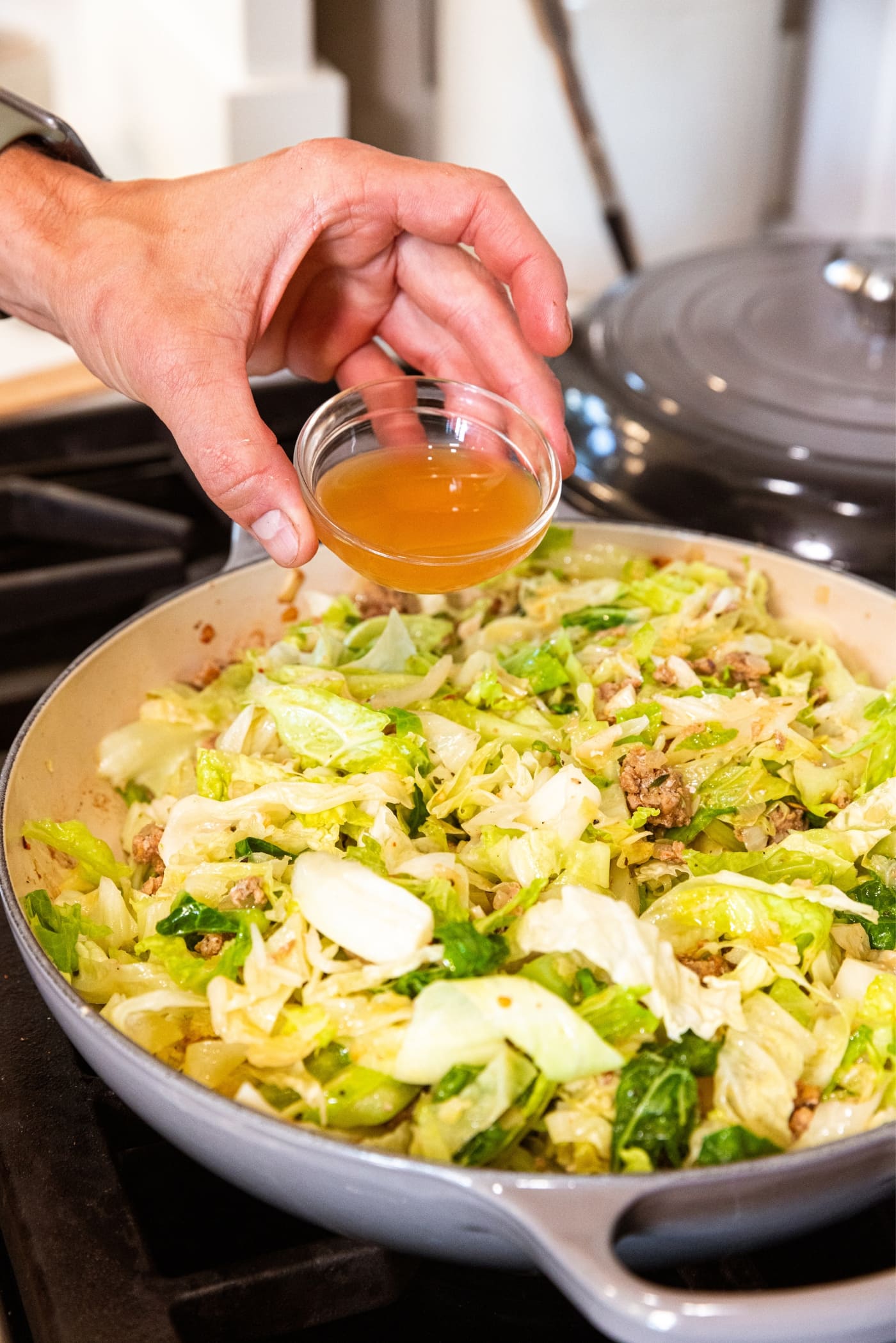 Hand holding small glass bowl filled with brown-colored apple cider vinegar about to pour it on top of sliced cabbage in large skillet.