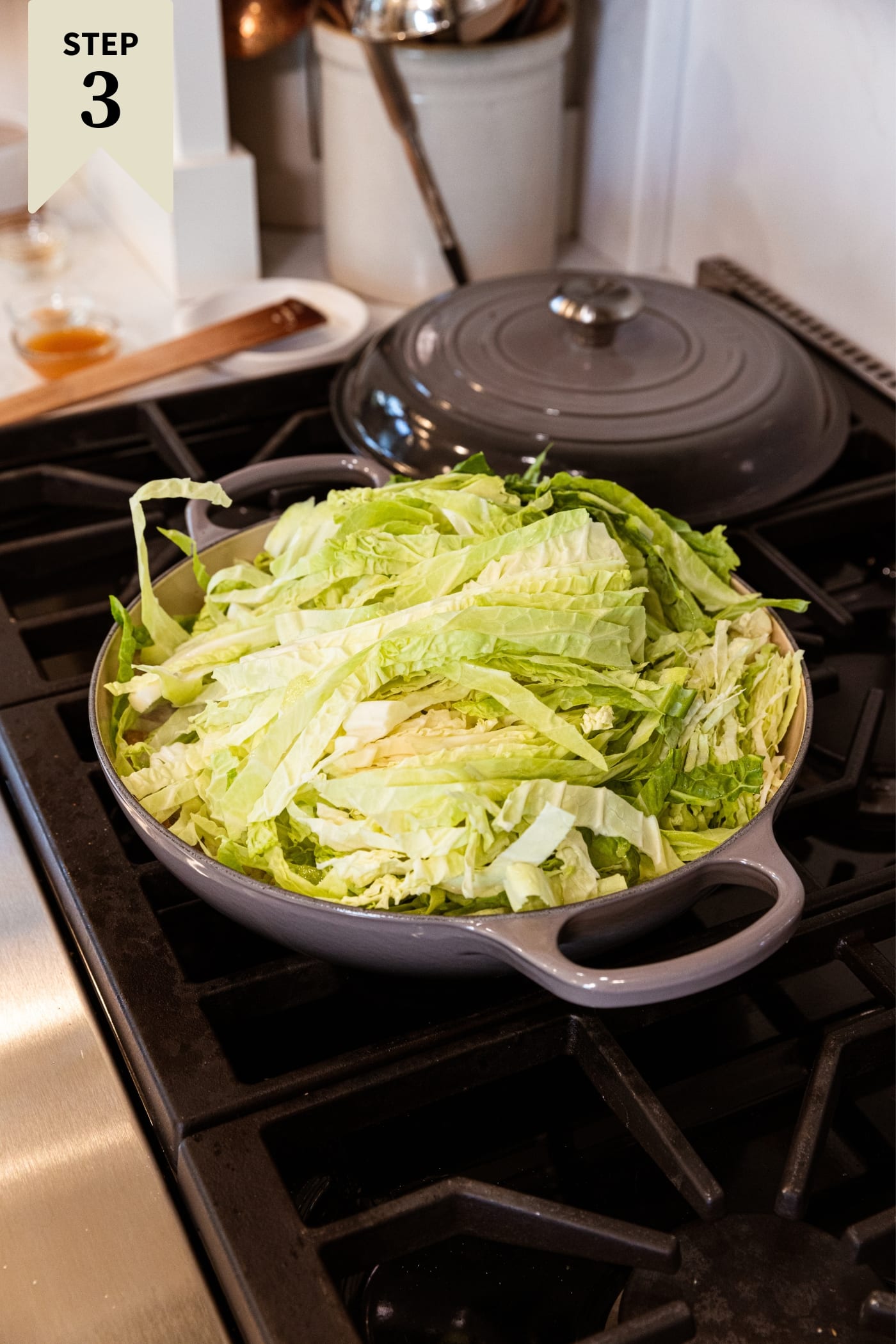 Large gray skillet on stovetop with lots of sliced cabbage beginning to wilt.