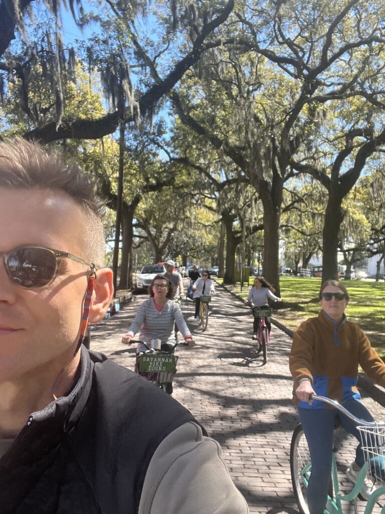 Kaleb Wyse riding bicycle with sunglasses on with family members riding bicycle behind him all under trees in Savannah Georgia.