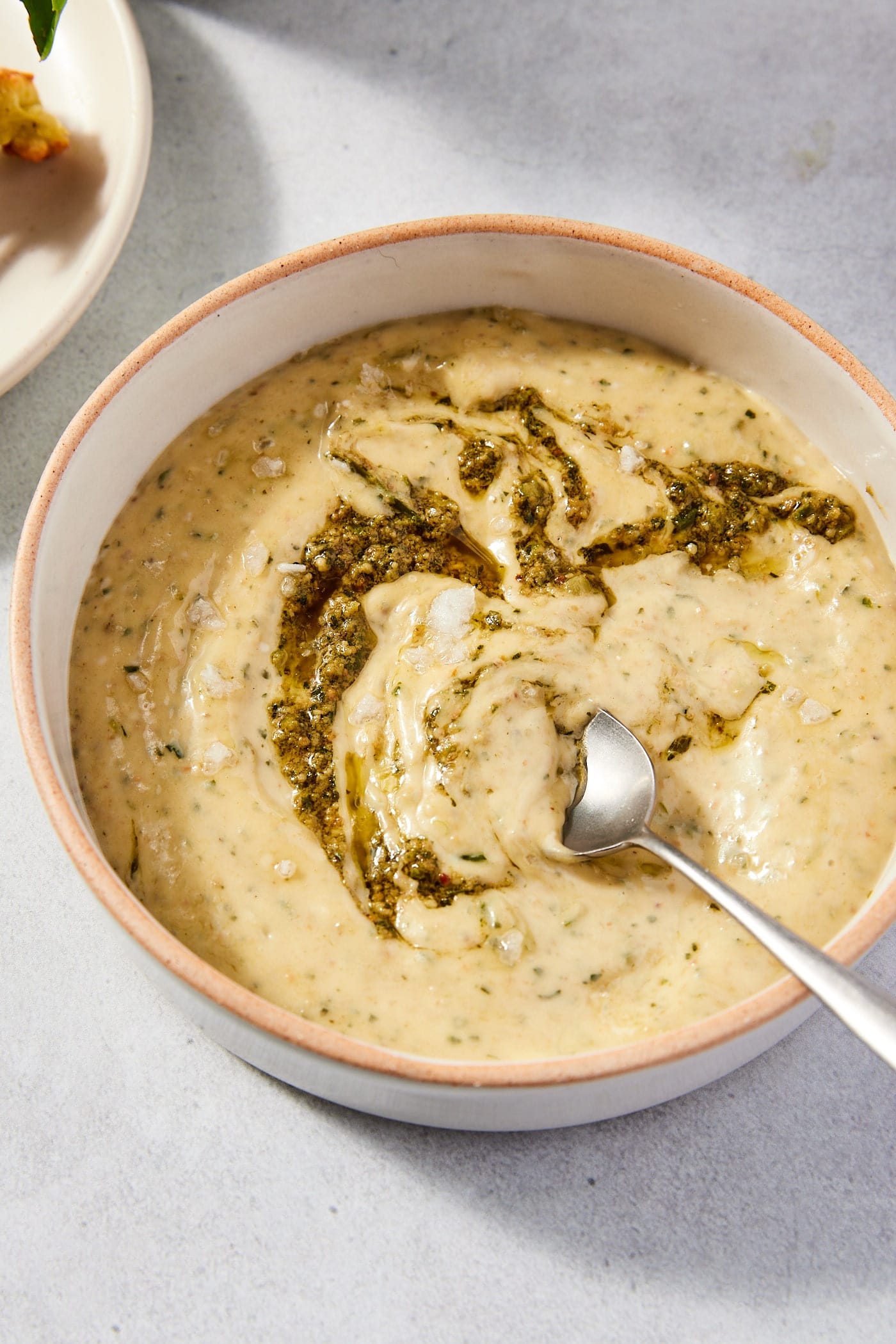 Stone surface with stoneware bowl filled with pesto aioli with pesto swirled over the top of the surface with spoon resting against bowl.