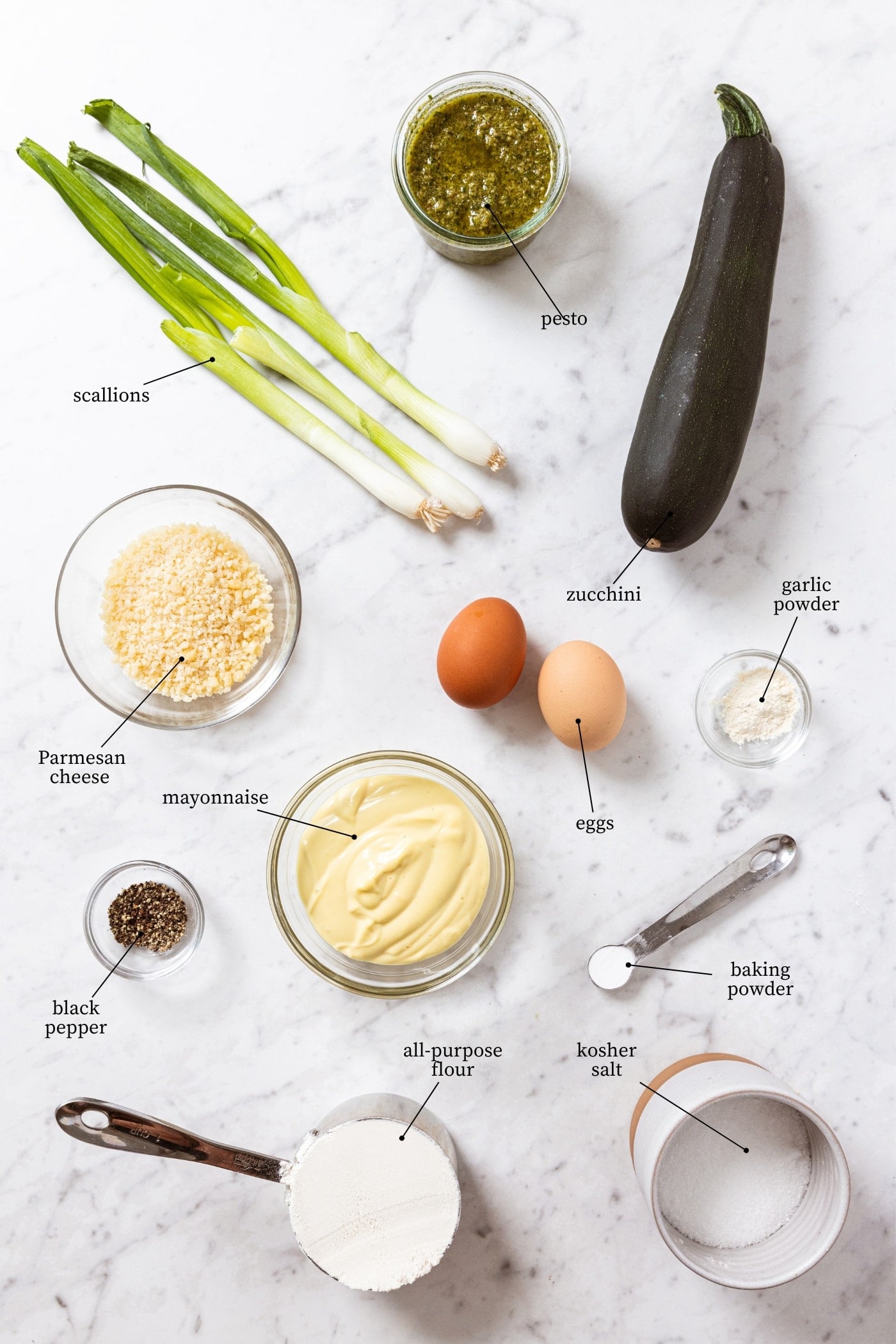 White marble surface filled with all ingredients needed to make zucchini fritters including zucchini, scallions, mayonnaise, eggs, spices, and more.