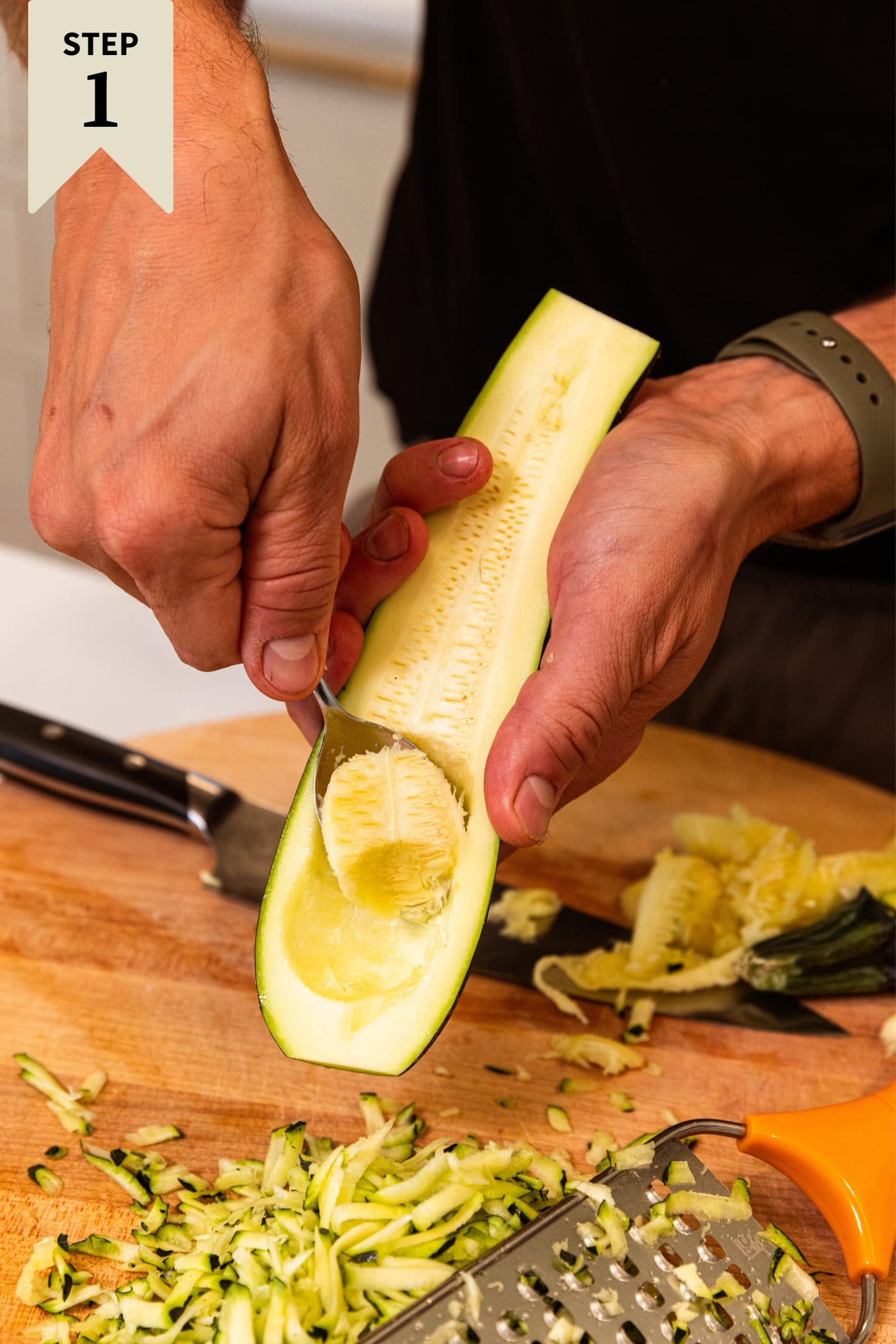 Hand holding halved zucchini scooping out the middle seeds with a spoon over a wood cutting board.