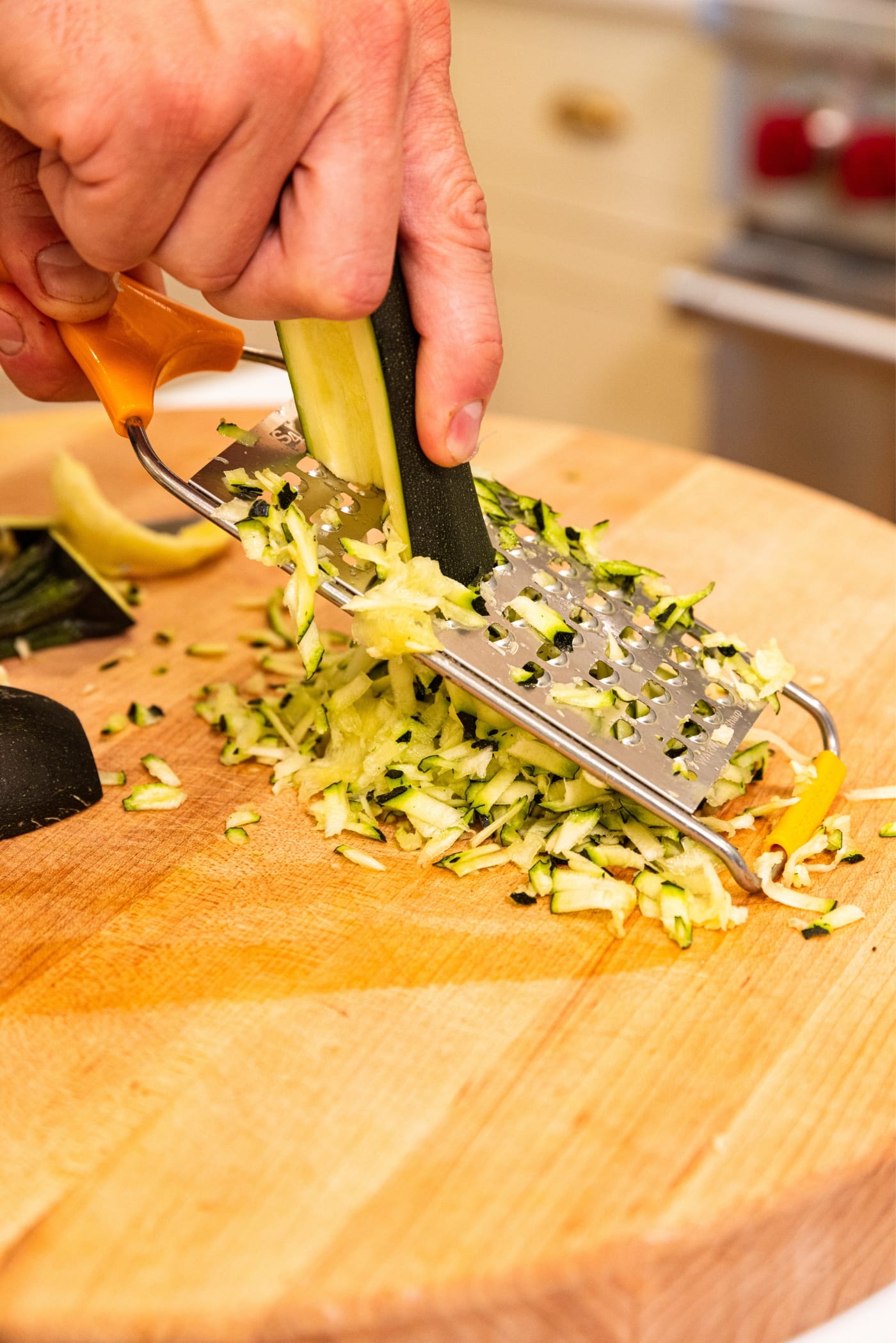 Hand holding zucchini sliced in half shredded it on grater on wood cutting board.