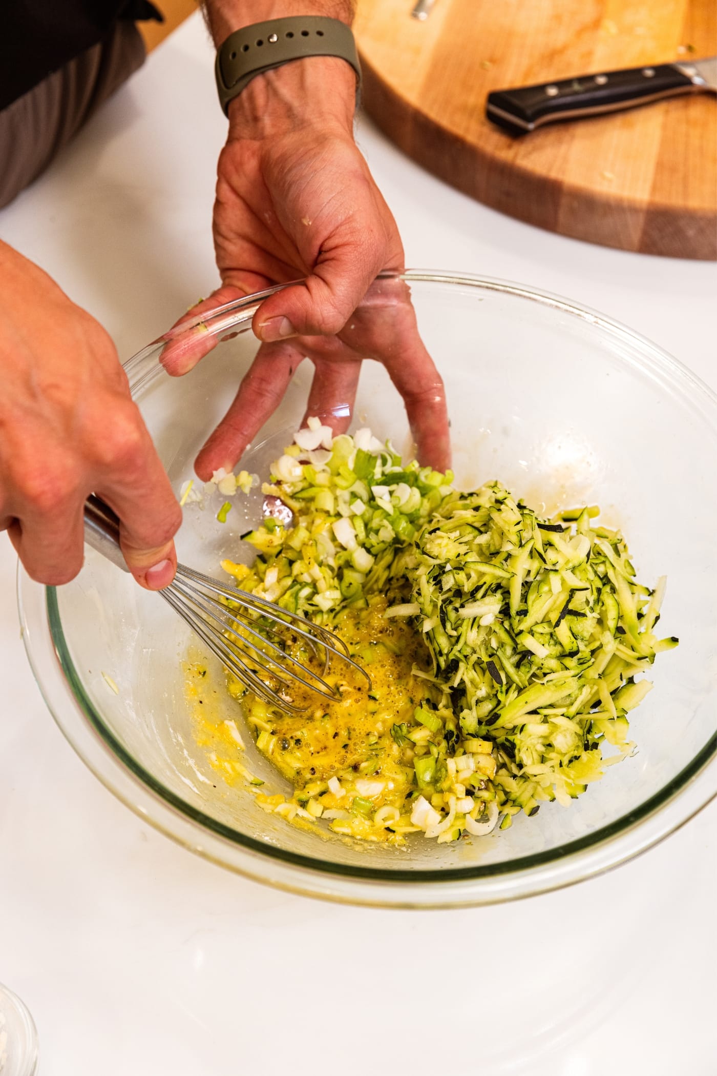 Hand holding metal whisk mixing together zucchini fritters batter in glass bowl on white surface.
