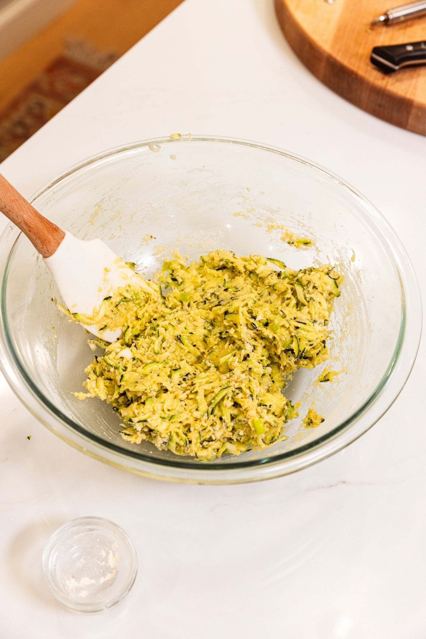 Glass bowl on white surface filled with zucchini fritters batter with spatula resting in bowl as well.