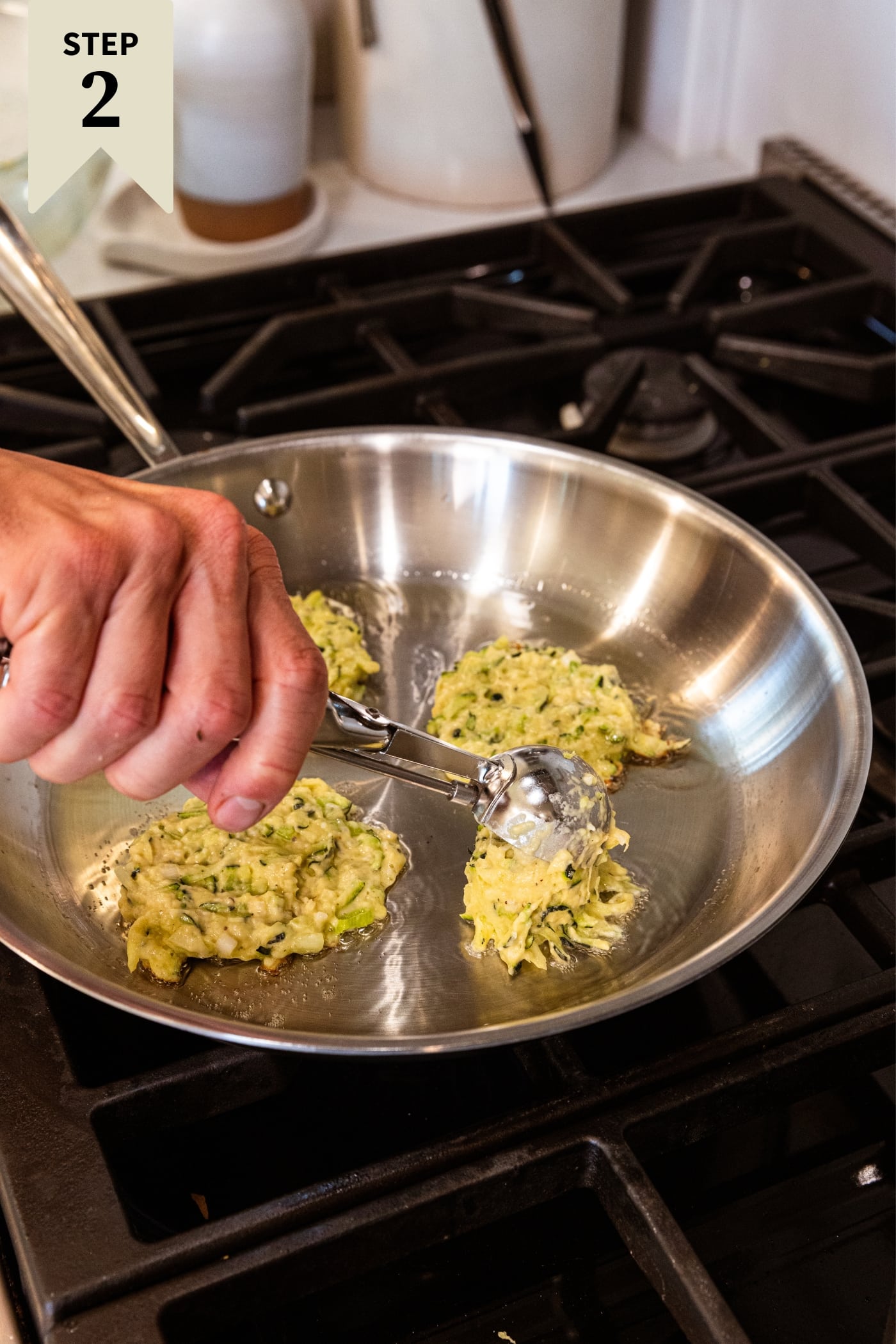 Hand using ice cream scoop to dollop zucchini fritters in metal skillet on stovetop.