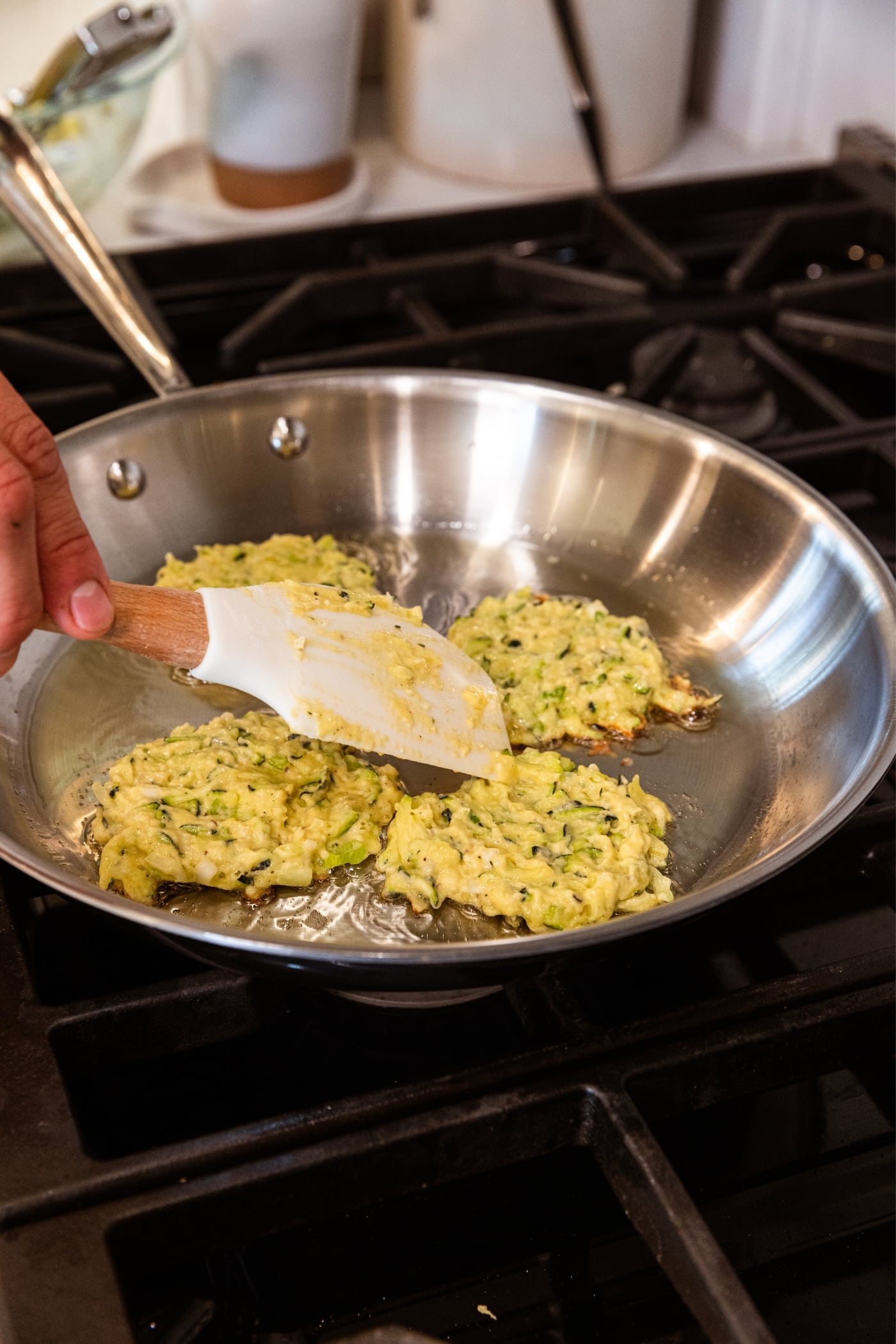 Hand using plastic spatula to spread out zucchini fritters in metal skillet on stovetop.