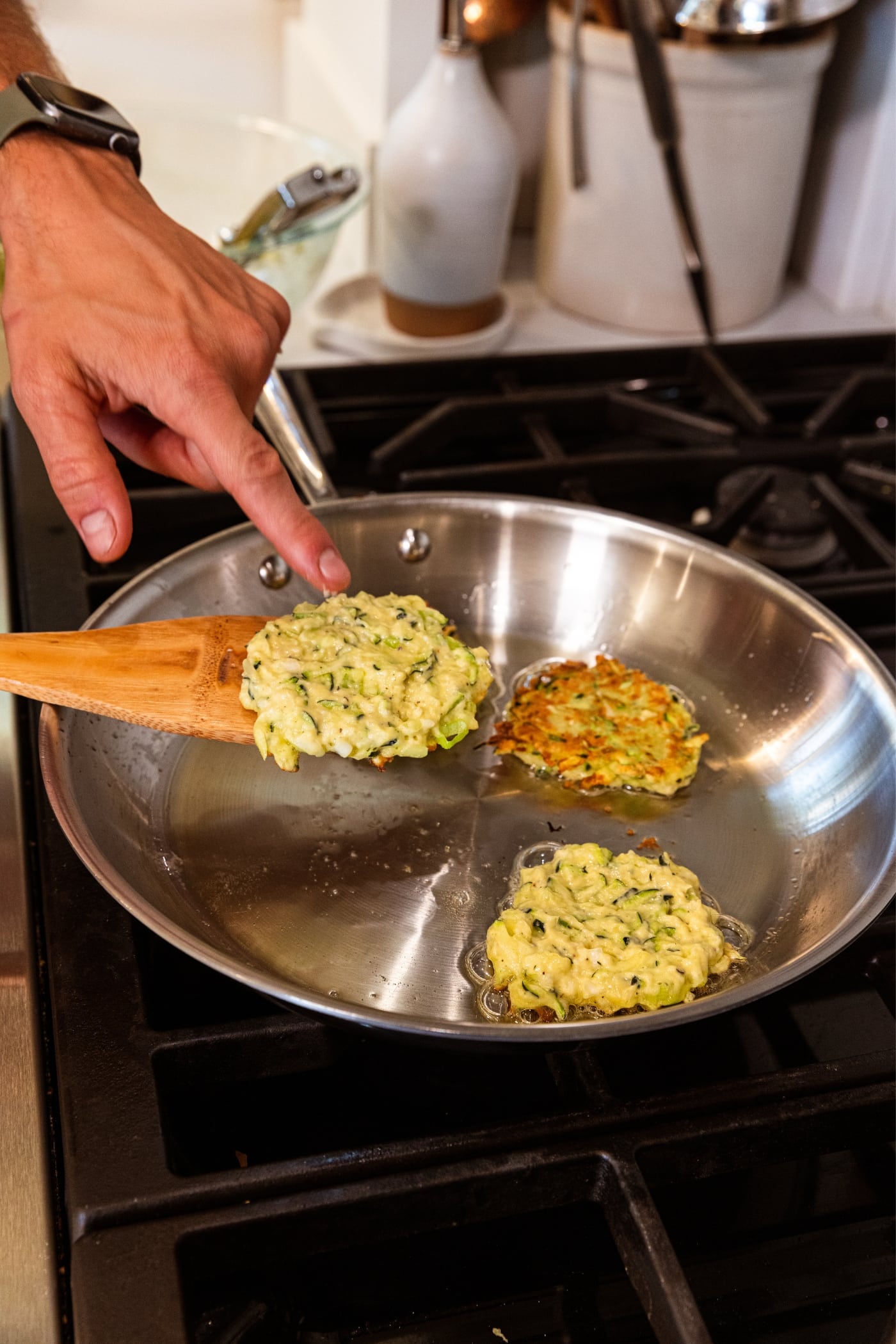 Hand using wooden spatula to flip cooking zucchini fritters in metal skillet on the stovetop.