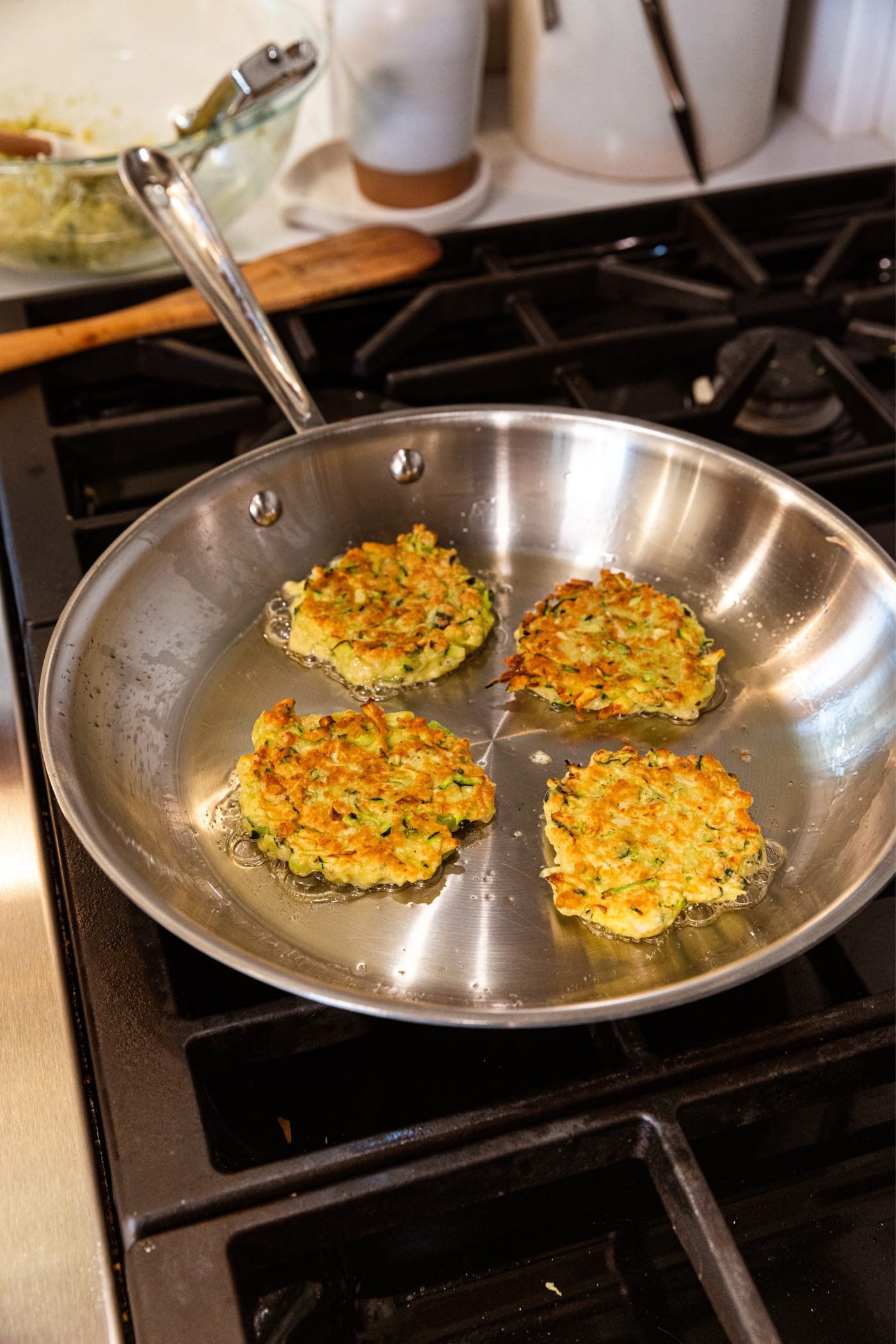 Metal skillet sitting on stovetop with browned cooking zucchini fritters just about done cooking in neutral oil.
