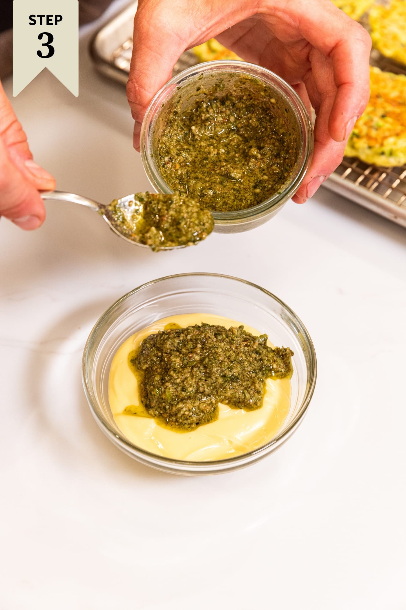 Hand holding glass bowl pouring basil pesto into a bowl filled with prepared mayonnaise all on white surface.