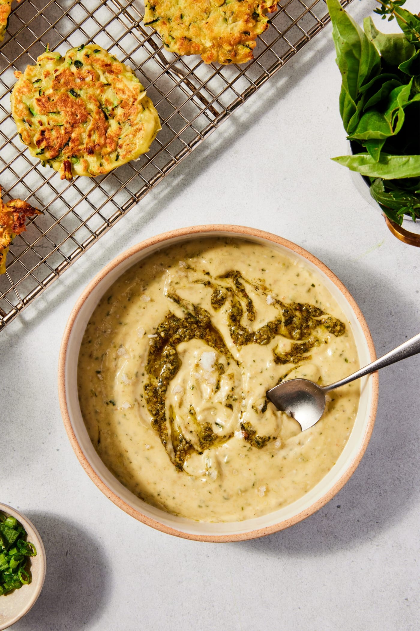 Stoneware bowl filled with pesto aioli with basil pesto swirled throughout sitting on stone surface with zucchini fritters cooling to the side.