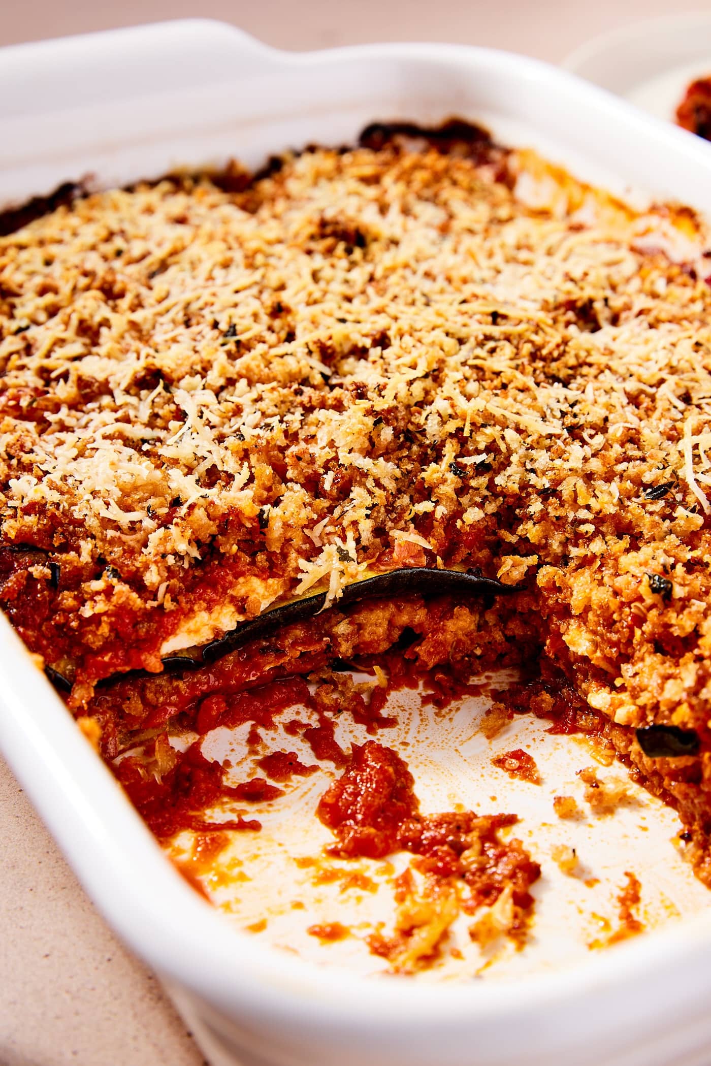 White rectangular baking dish filled with zucchini parmesan showing interior layers of tomato sauce, cheese, and breadcrumbs.