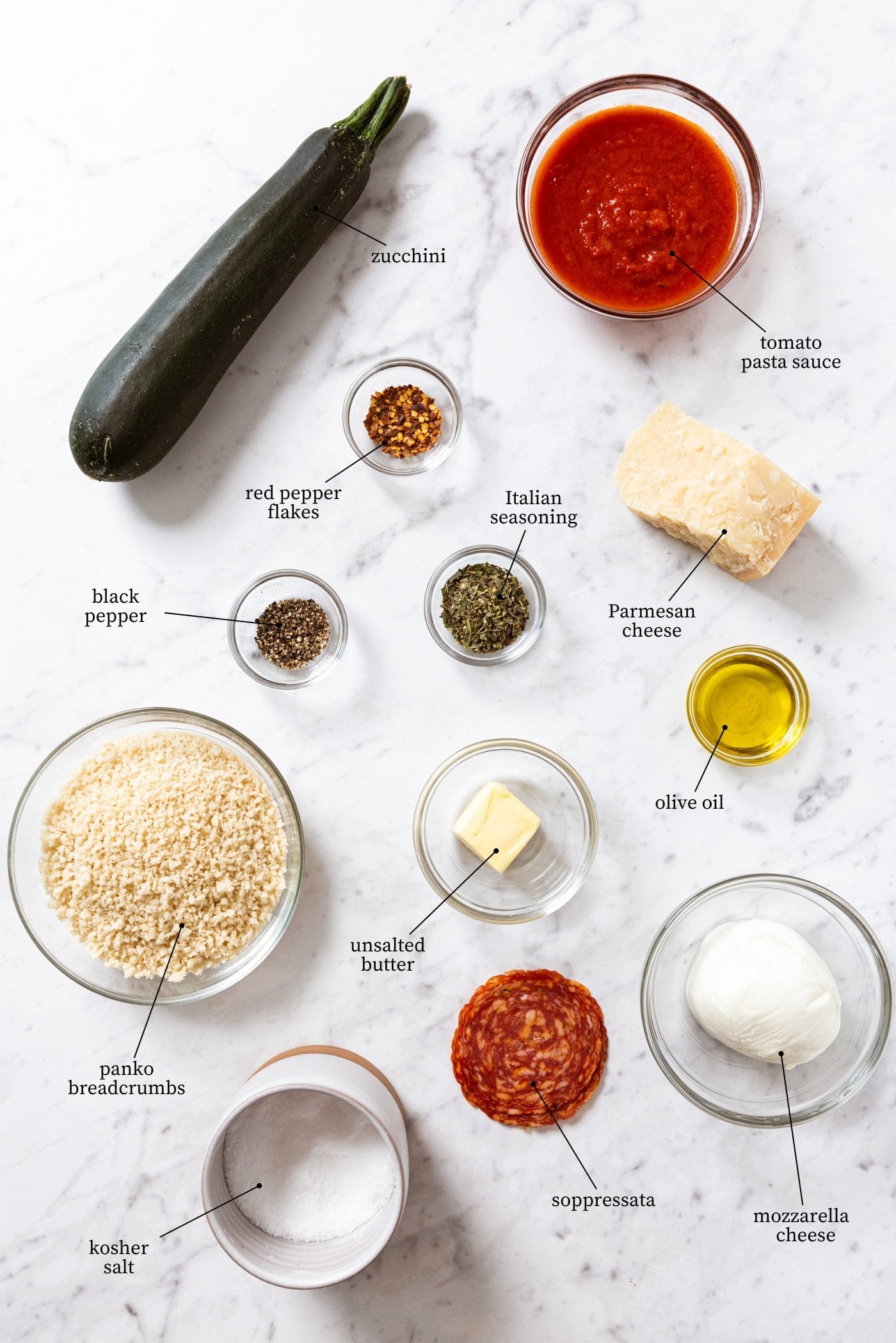 White marble surface filled with all ingredients needed to make zucchini parmesan including breadcrumbs, tomato sauce, zucchini, meat, spices, and more.