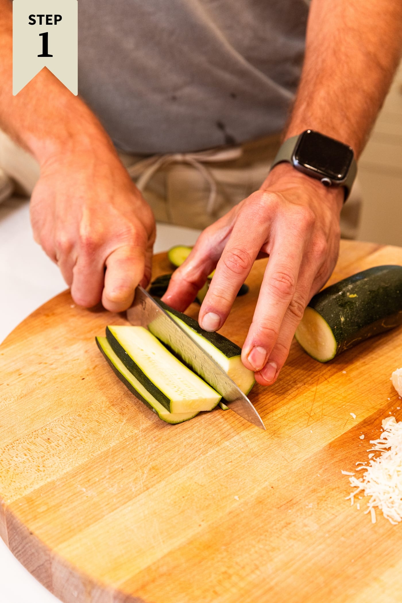Hands slicing zucchini into planks on wooden cutting board with more zucchini to the side.