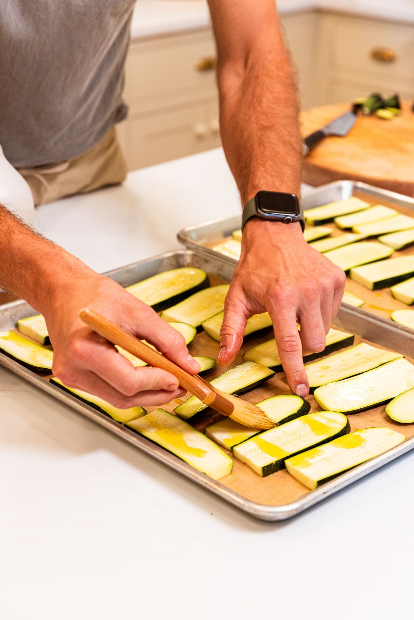 Hand using brush to spread out olive oil on planks of zucchini sitting on baking dish on white surface.