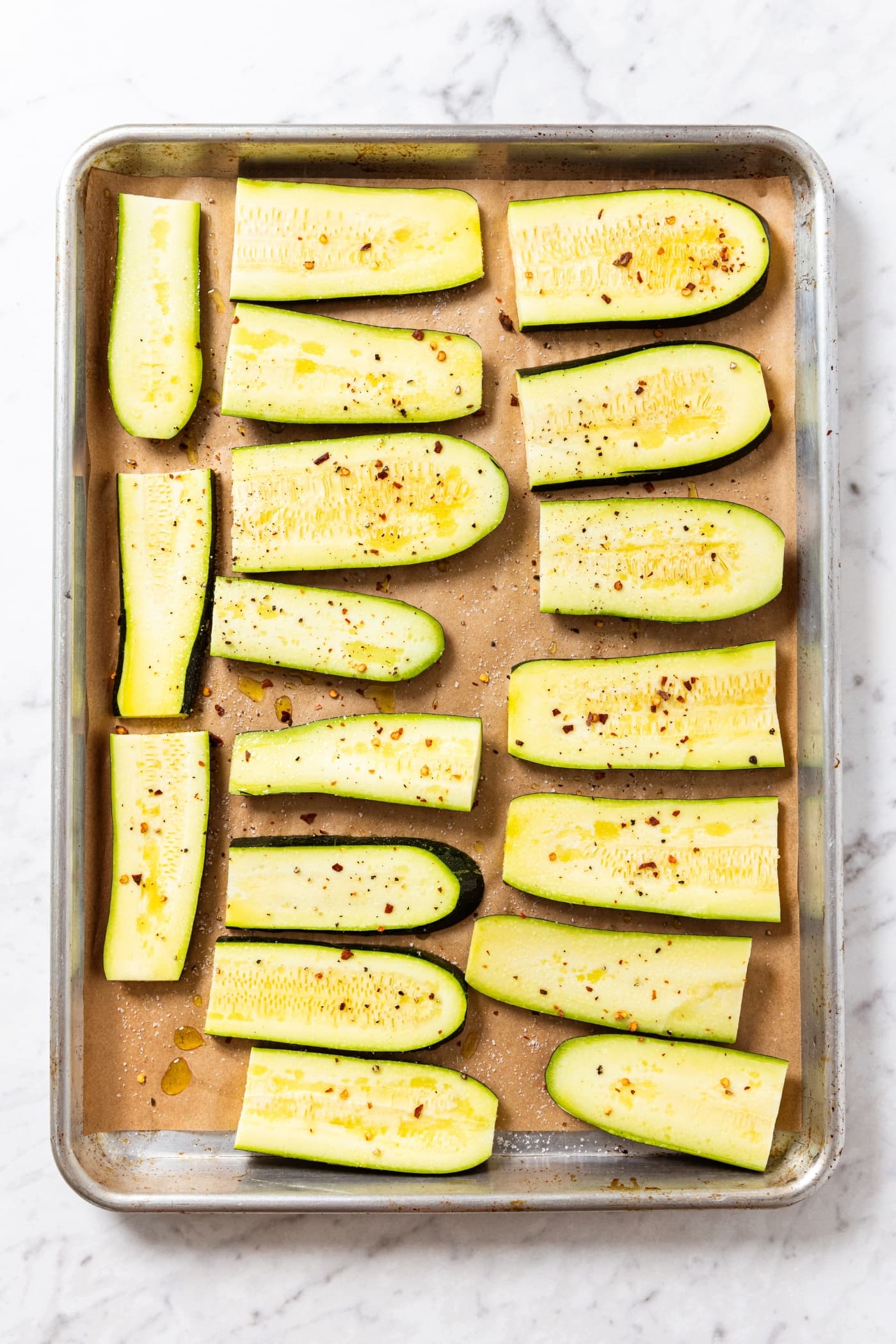 Roasting pan filled with slices of zucchini sprinkled with salt, pepper, and olive oil right before going into the oven to bake.