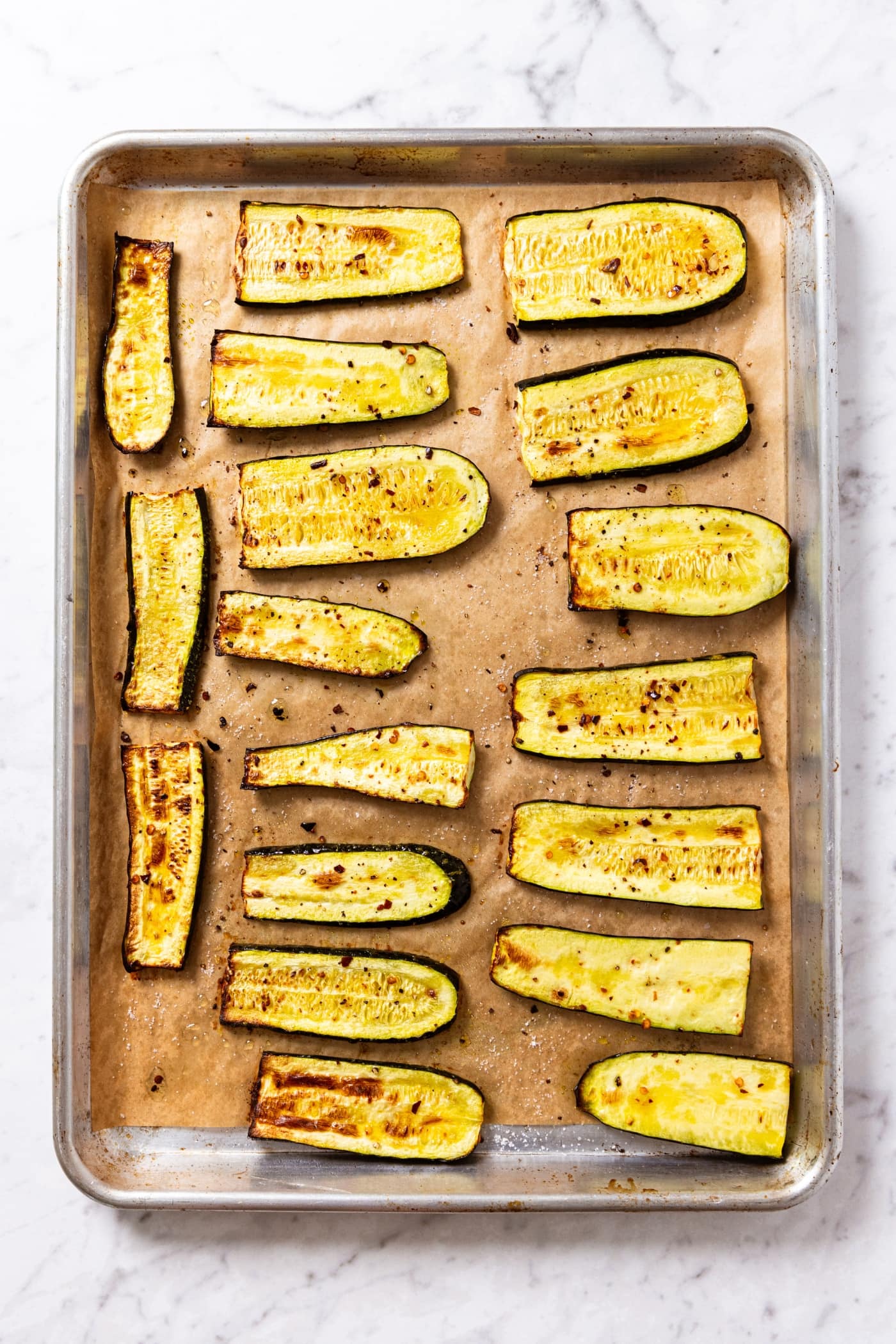 Roasted zucchini slices on baking sheet after coming out of the oven sitting on white marble surface.
