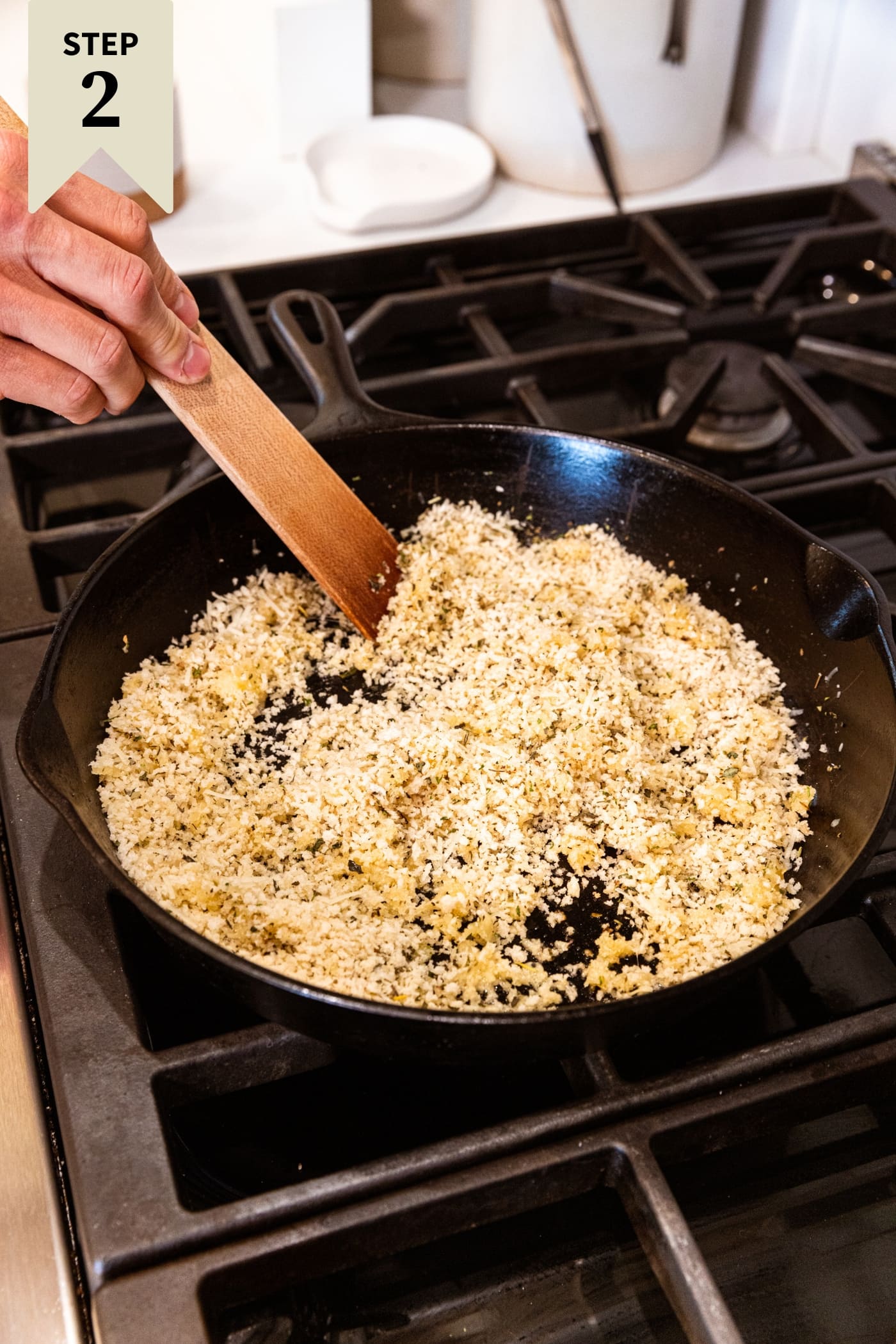 Hand holding wooden spatula mixing around uncooked breadcrumbs in black cast iron skillet on stovetop.