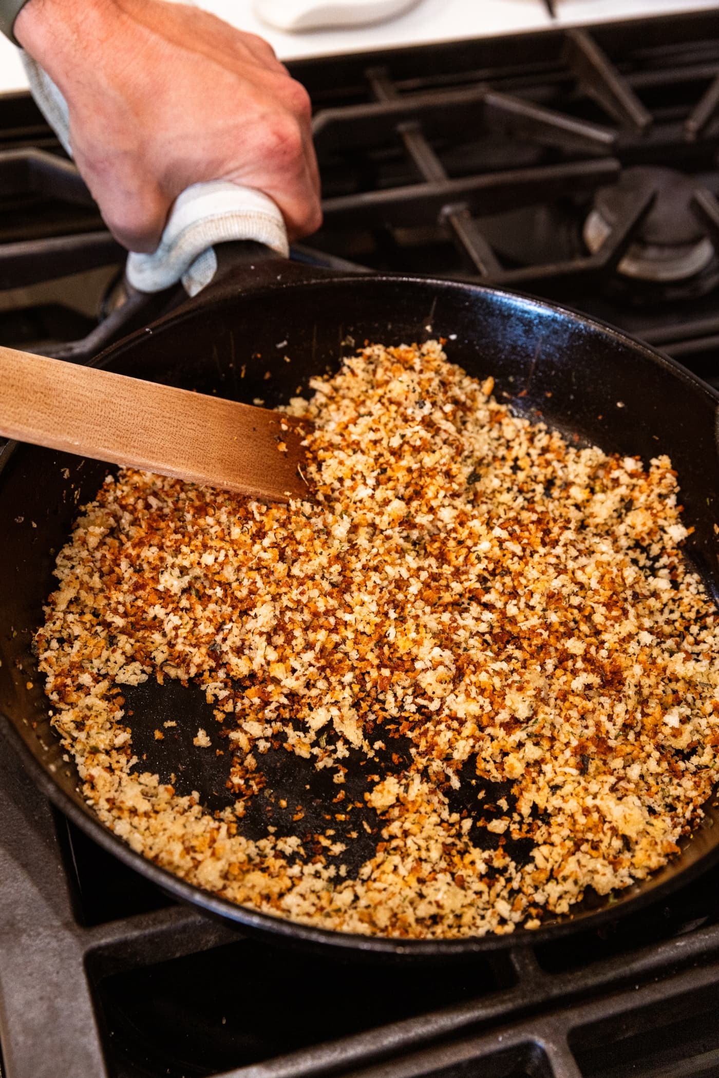 Wooden spatula stirring together cooked and browned breadcrumbs in black wooden skillet on stovetop.