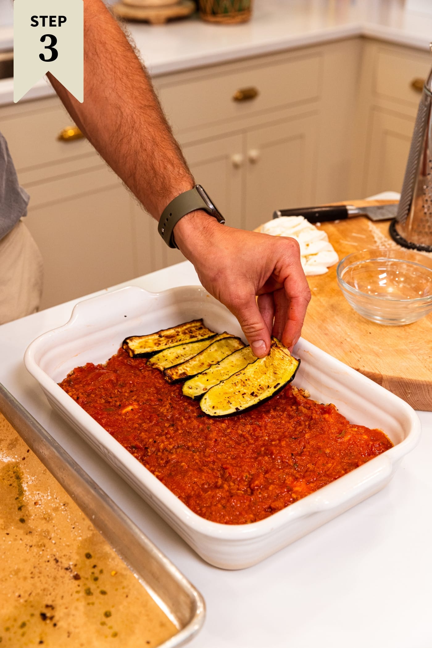 White baking dish with layer of tomato sauce with hand placing slices of roasted zucchini on top.