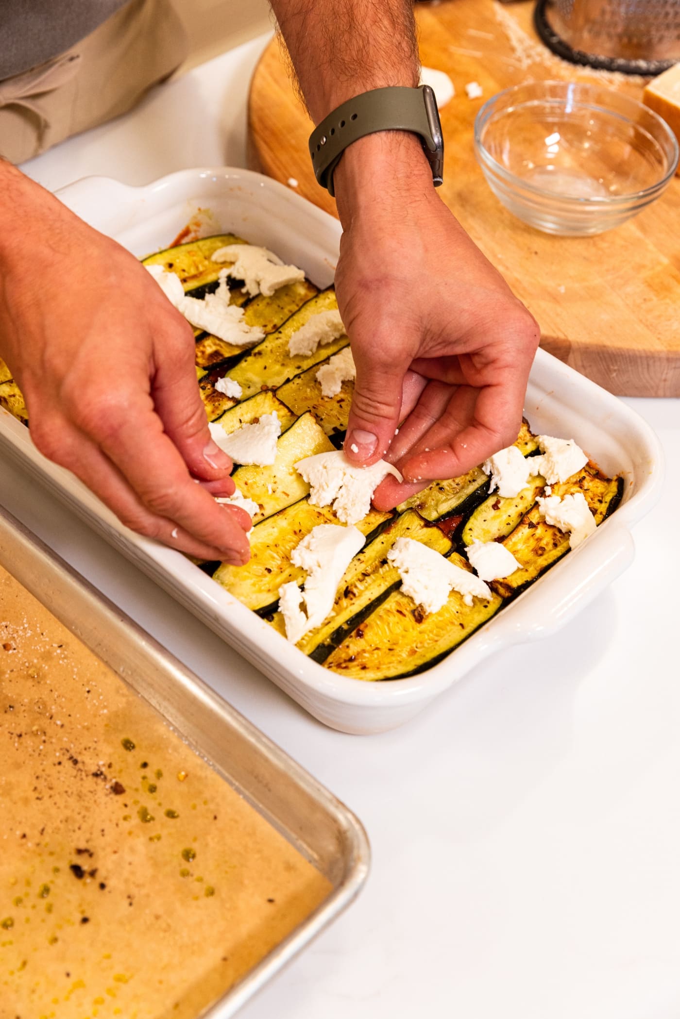 Hands placing mozzarella cheese on top of layer of roasted zucchini slices in white baking dish on white surface.