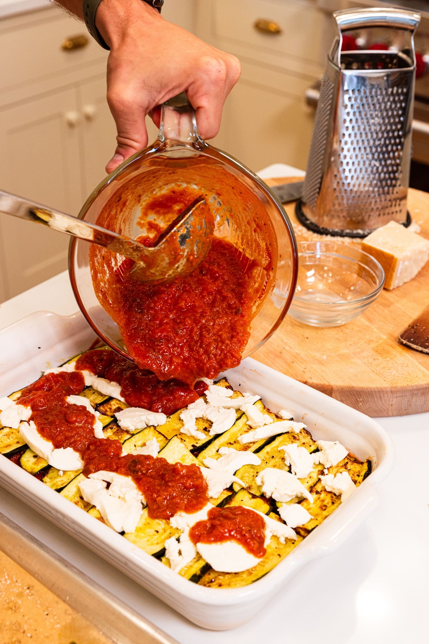 Glass measuring cup pouring tomato sauce on top of layer of mozzarella cheese and roasted zucchini pieces.