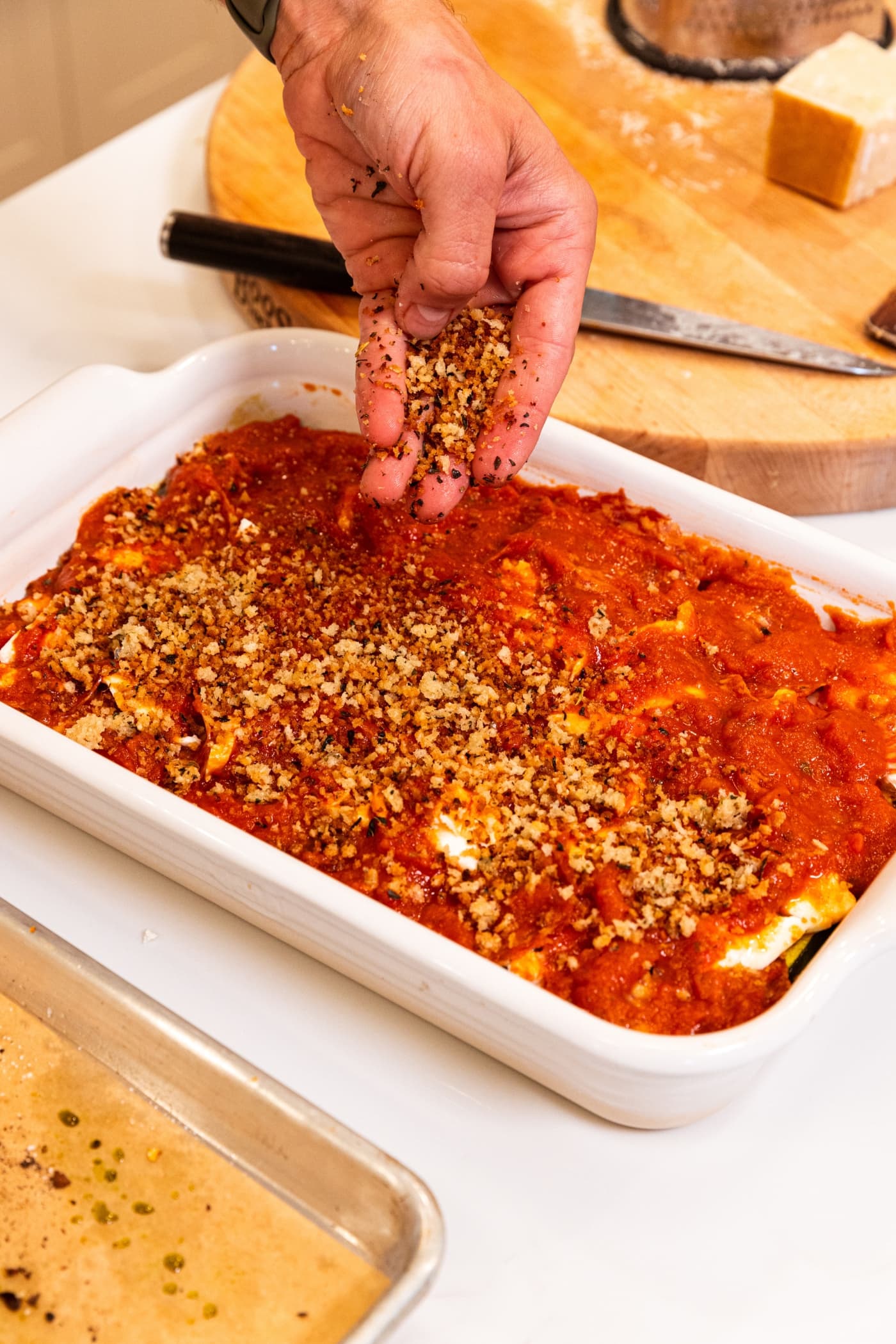 Hand spreading breadcrumbs on top of layer of tomato sauce in white baking dish on white surface.
