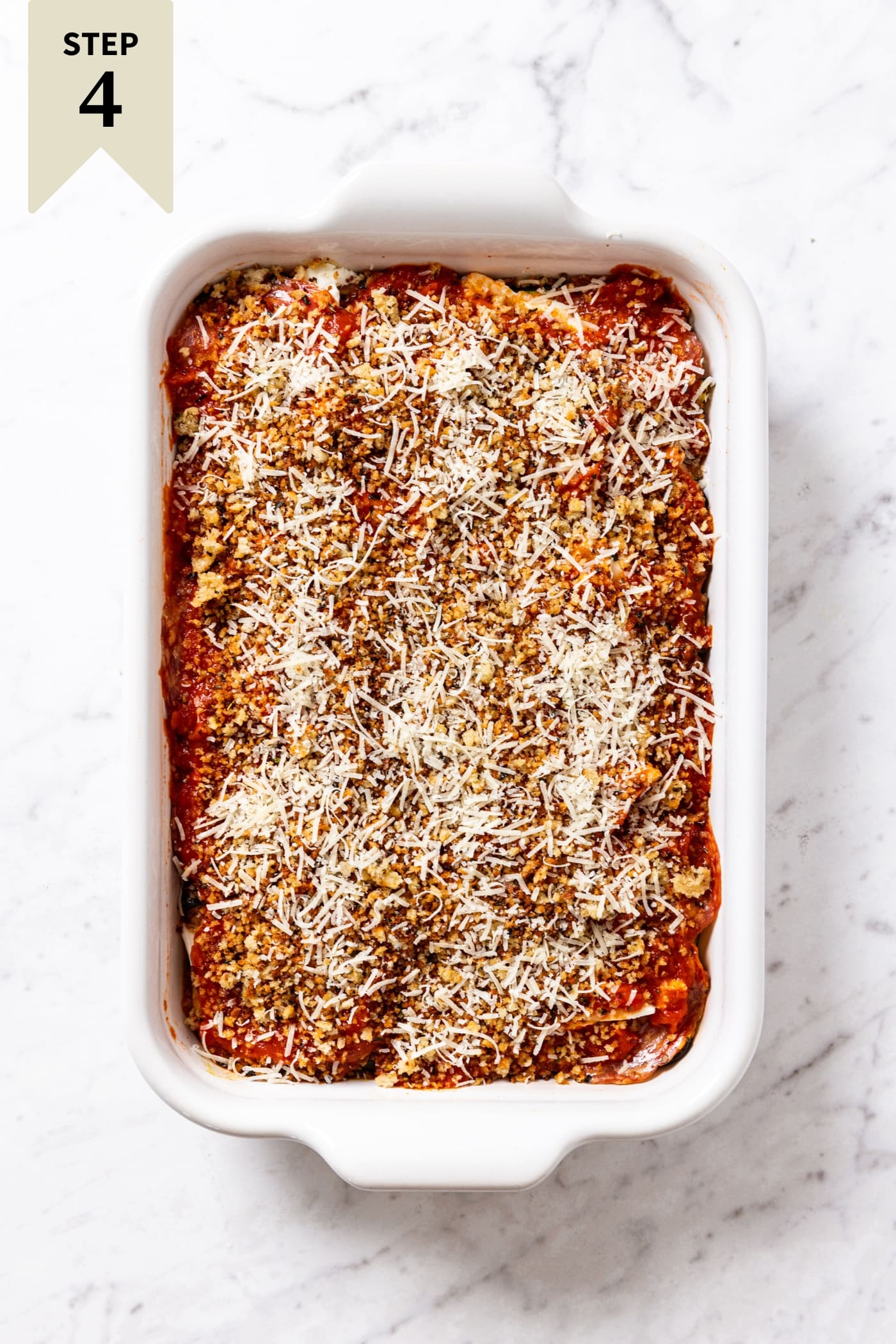 White marble surface with white baking dish filled with unbaked zucchini parmesan with shredded Parmesan cheese and breadcrumbs all over the top of tomato layer.