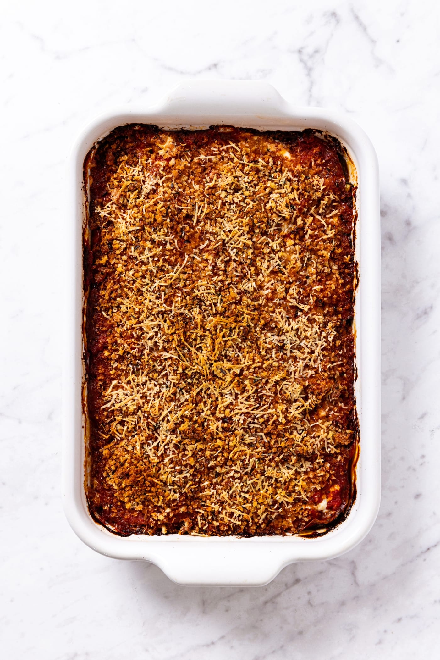 White baking dish filled with baked zucchini parmesan recipe sitting on white marble surface.