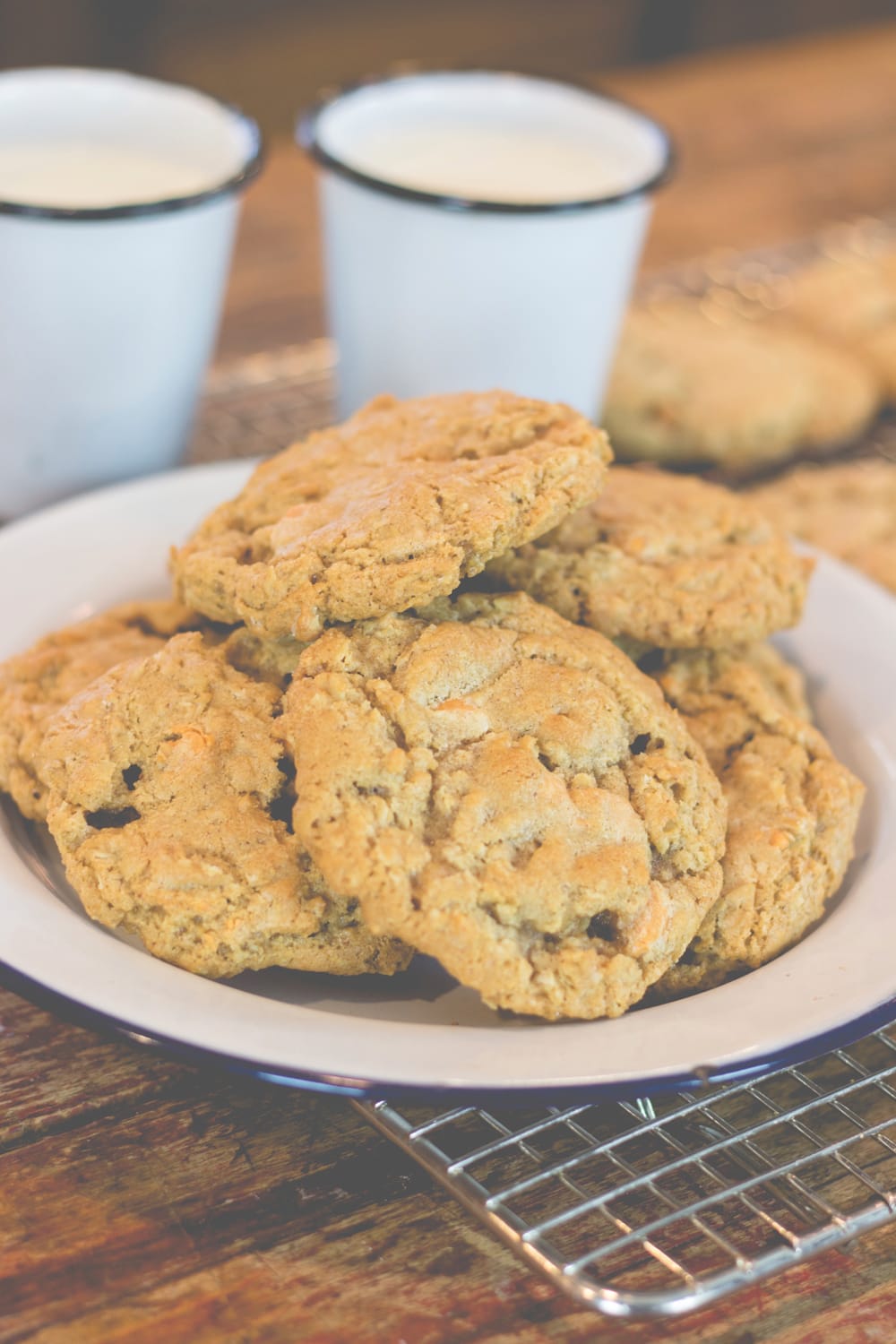 A stack of cookies sits on a plate with glasses of milk behind.