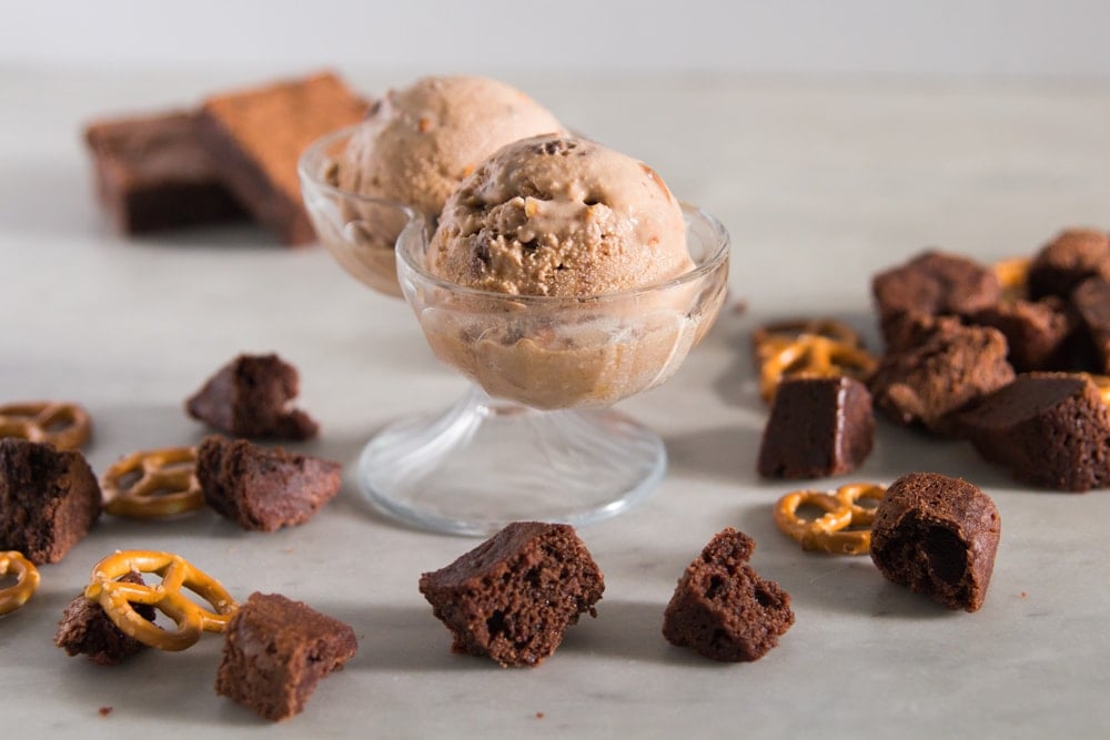 Two scoops of ice cream sit in a glass container with brownies and pretzels scattered beneath.