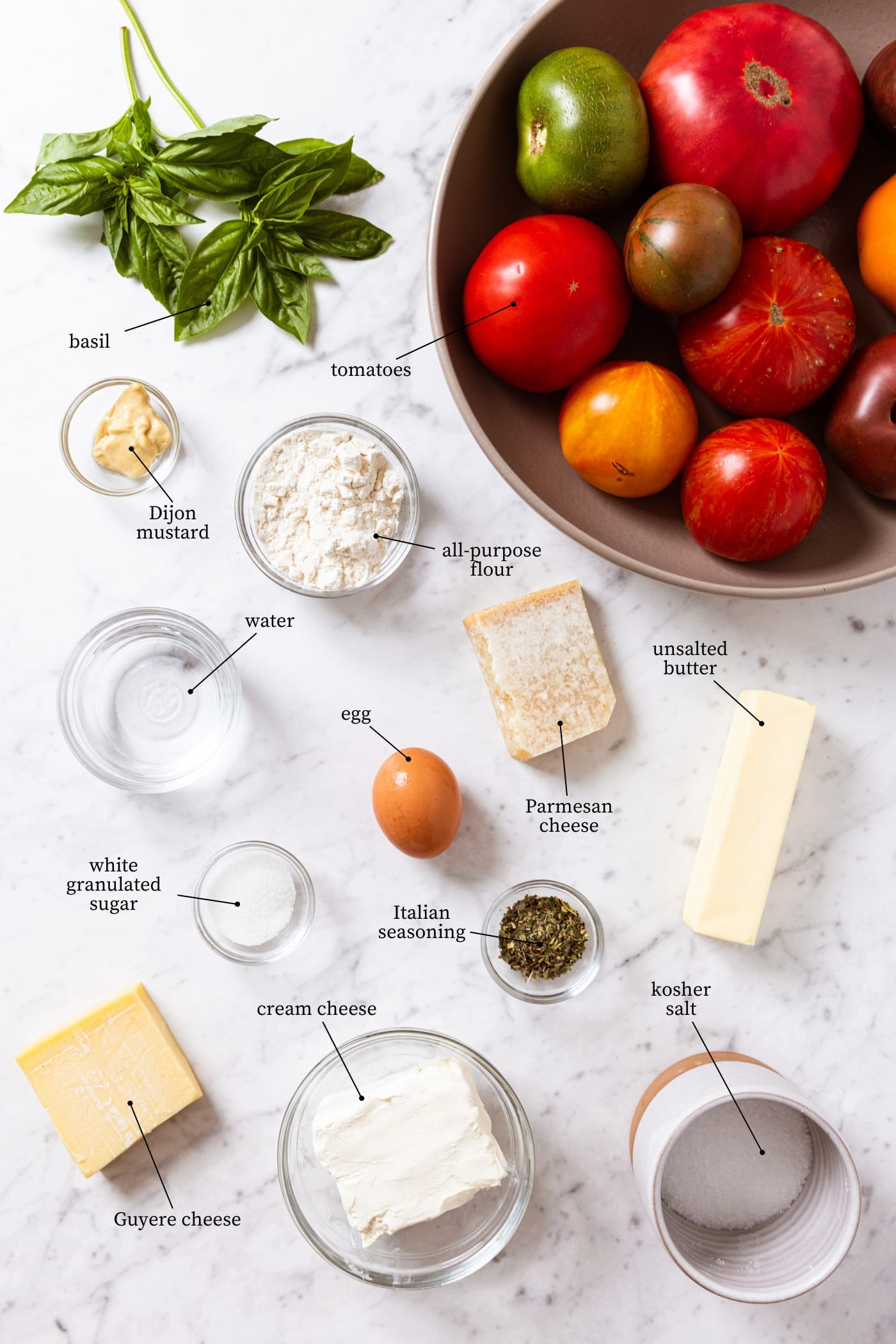 White marble surface filled with all ingredients needed to make a fresh tomato galette including tomatoes, basil, flour, egg, cheese, and more.