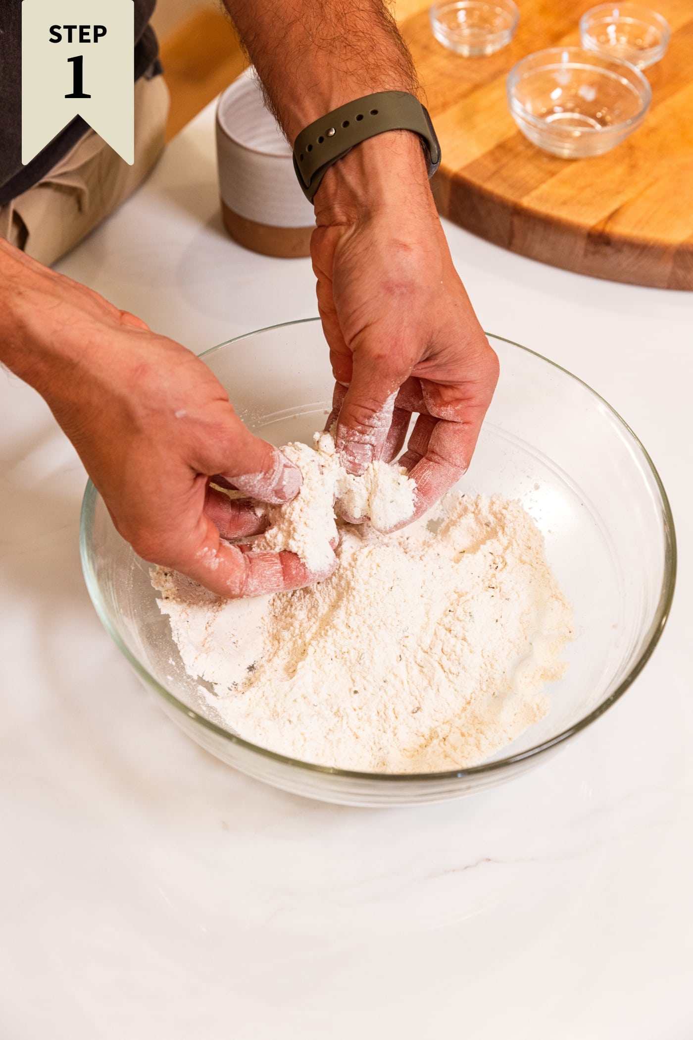 Two hands mixing together flour in a glass bowl on white surface with wood cutting board in background.
