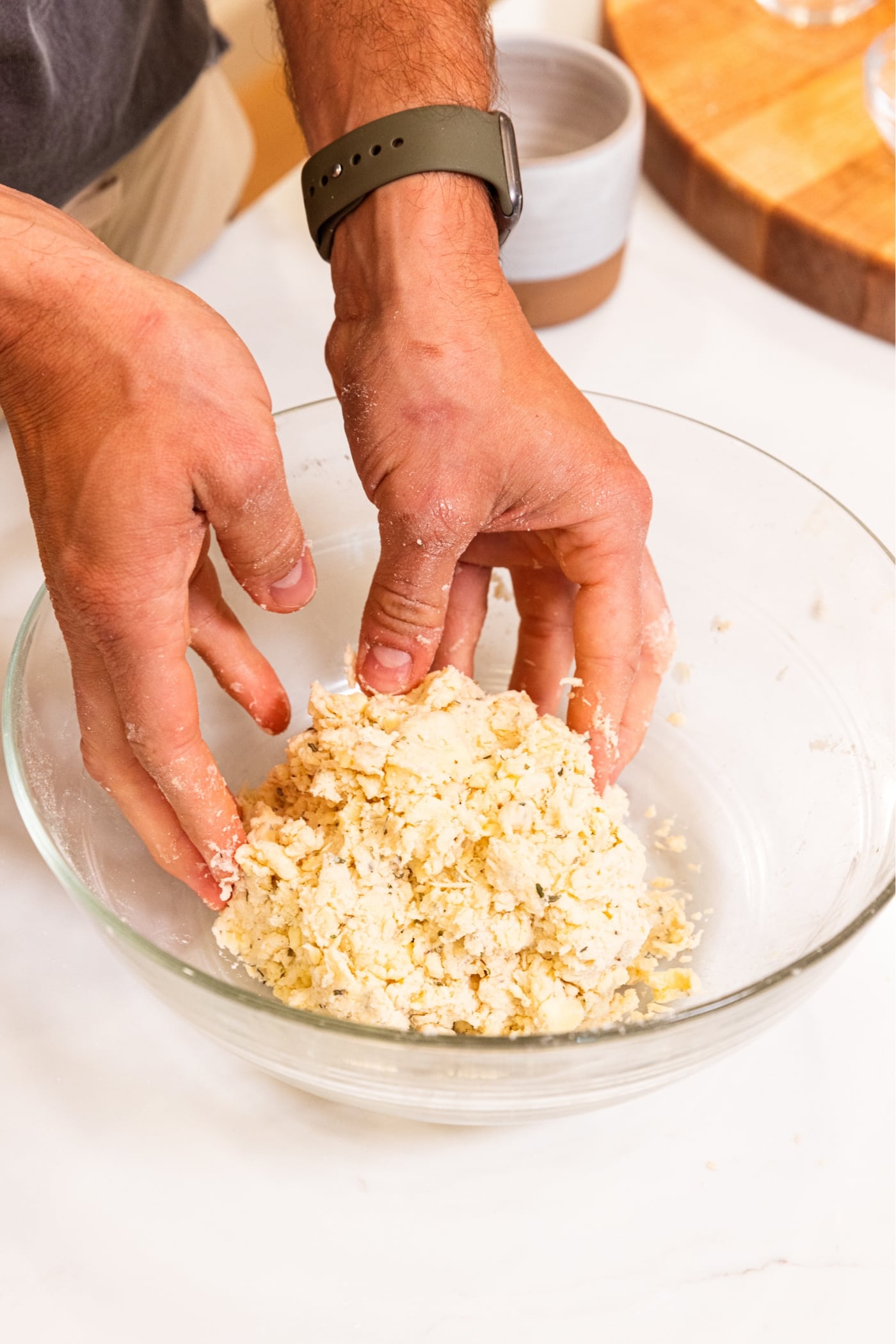 Two hands pushing together formed dough in a large glass bowl with pieces of dried herbs showing.