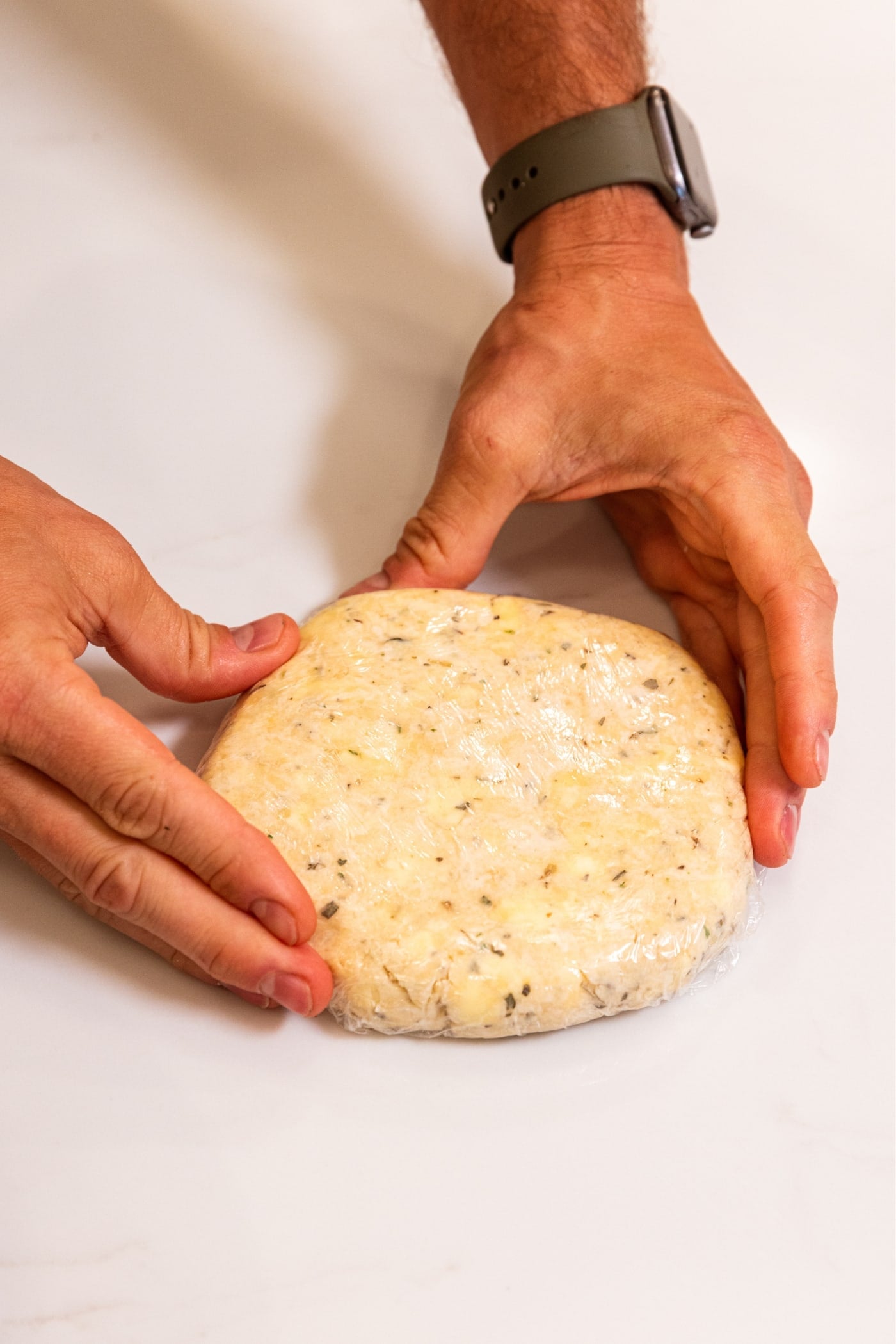 Hands holding a round of dough wrapped in plastic wrap sitting on white surface.