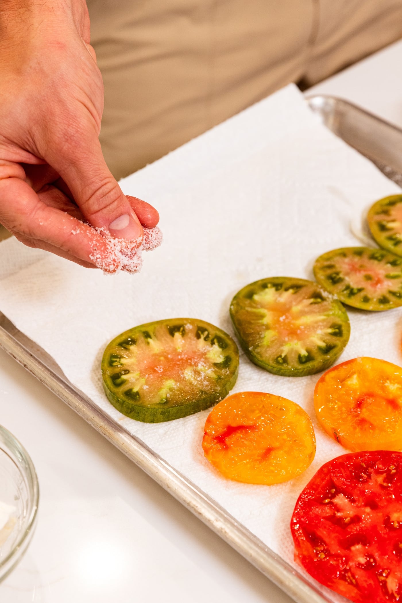 Hand sprinkling kosher salt on top of slices of tomato on paper towel-lined baking sheet.