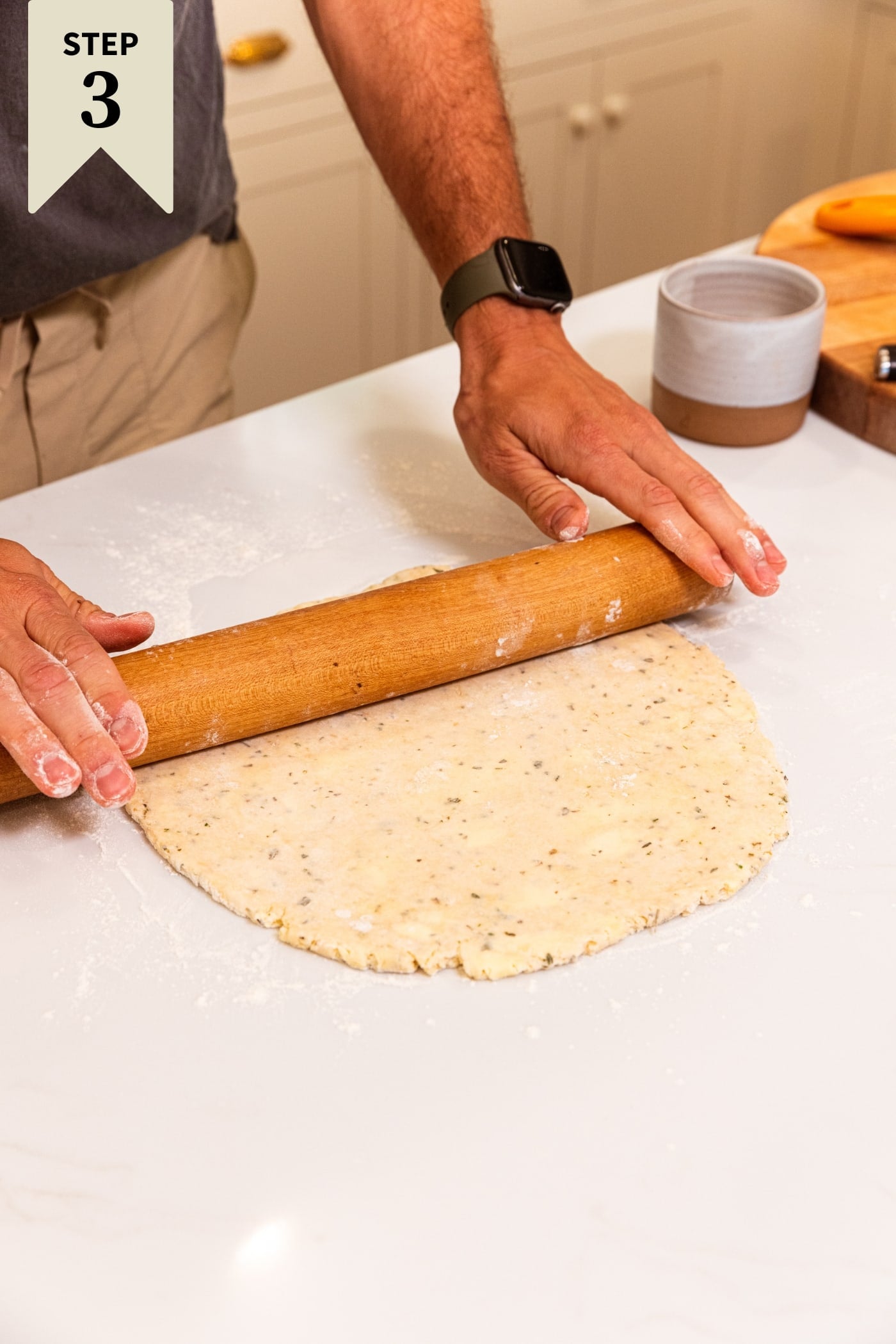 Hands holding wooden rolling pin rolling out dough on a white surface.