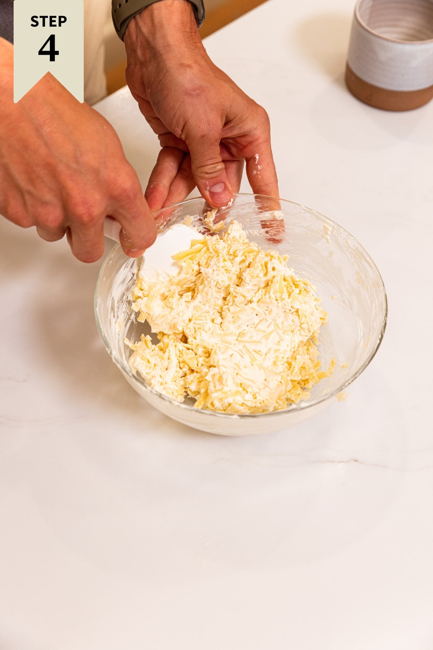 Hand holding spatula mixing together cream cheese and shredded cheese in small glass bowl on white surface.