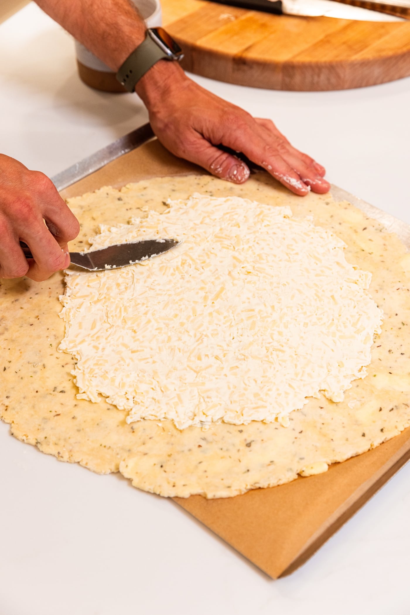 Hand using offset spatula to spread cream cheese mixture on top of unbaked dough on baking sheet.