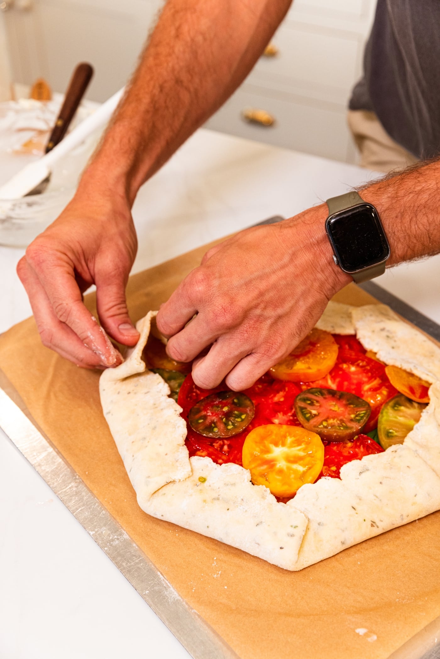 Hand pressing dough for a tomato galette with fresh slices of tomato all on baking sheet.