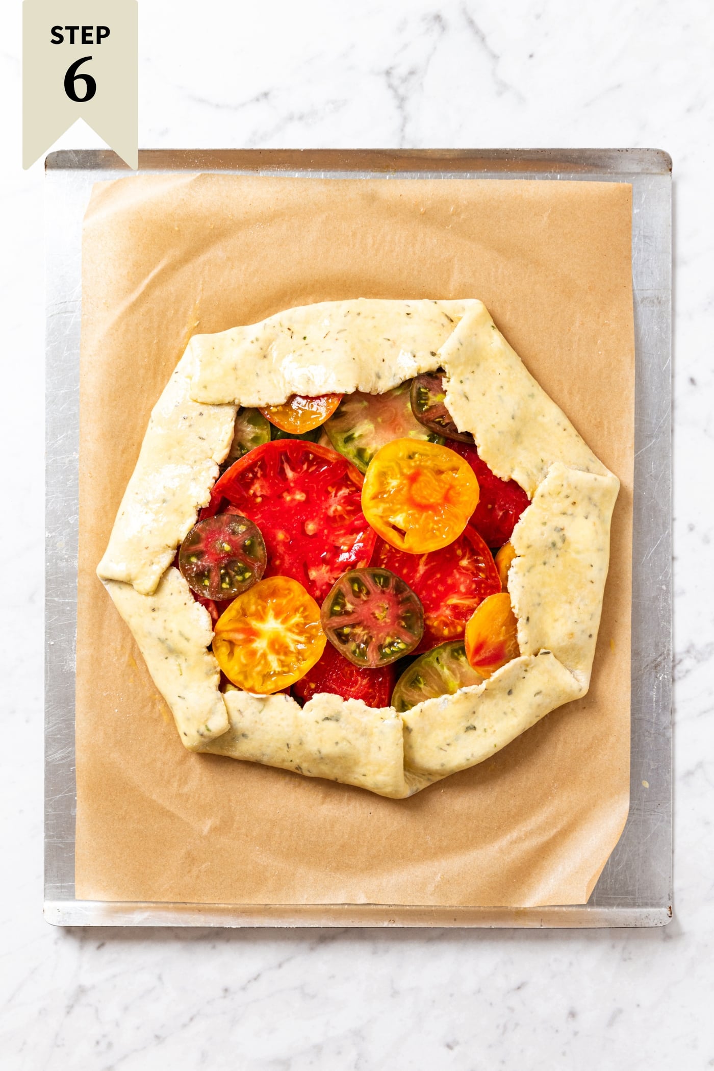 Unbaked tomato galette sitting on piece of brown parchment on a baking sheet before going into the oven to bake.